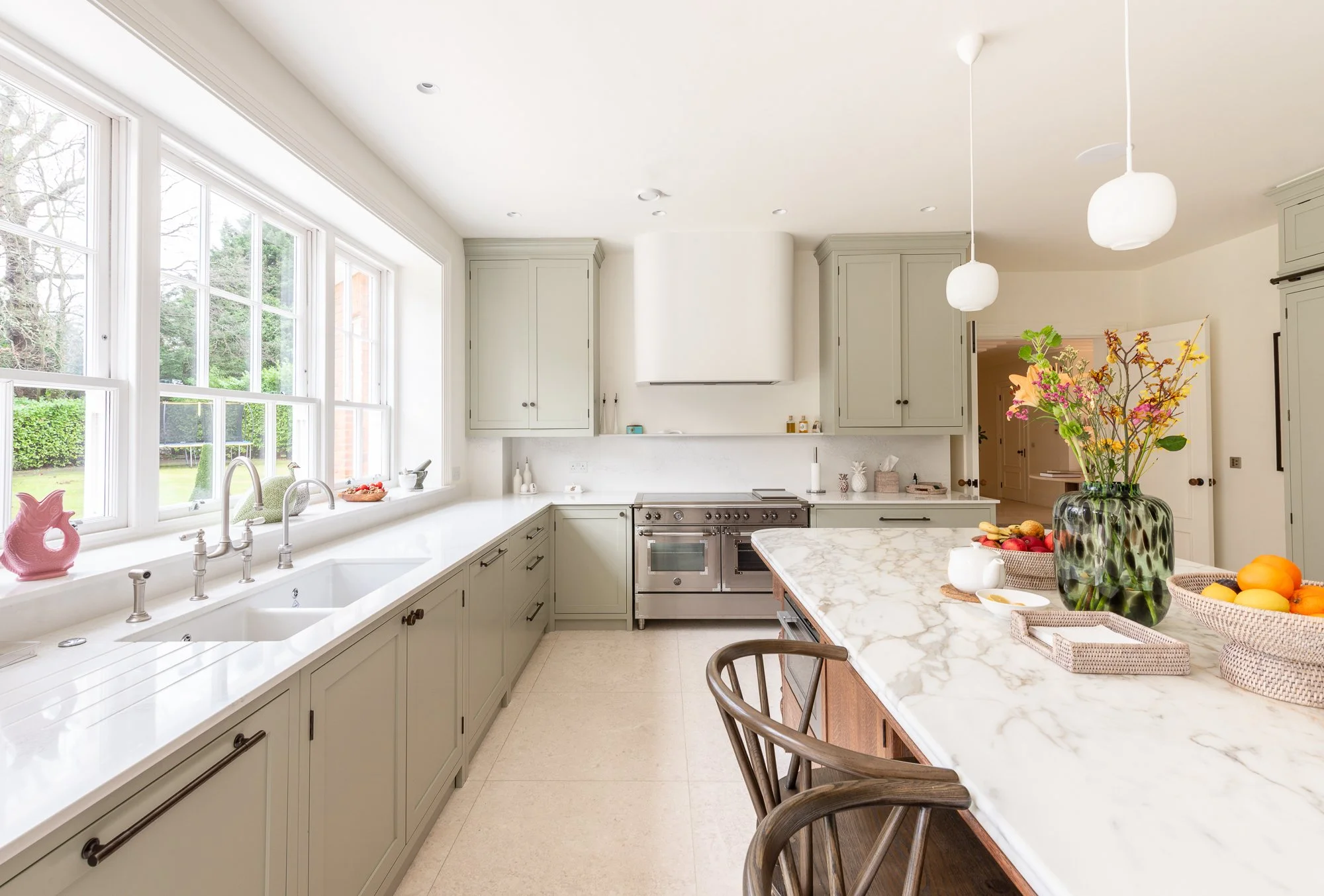 Bright kitchen with large windows, white cabinetry, marble countertop, stainless steel oven, and decorative fruits and flowers on the island.