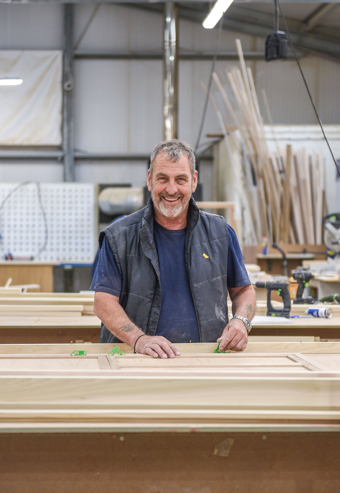 A man with gray hair and a beard, smiling, working on woodworking projects in a workshop. The workshop has tools, wood pieces, and partially assembled furniture.