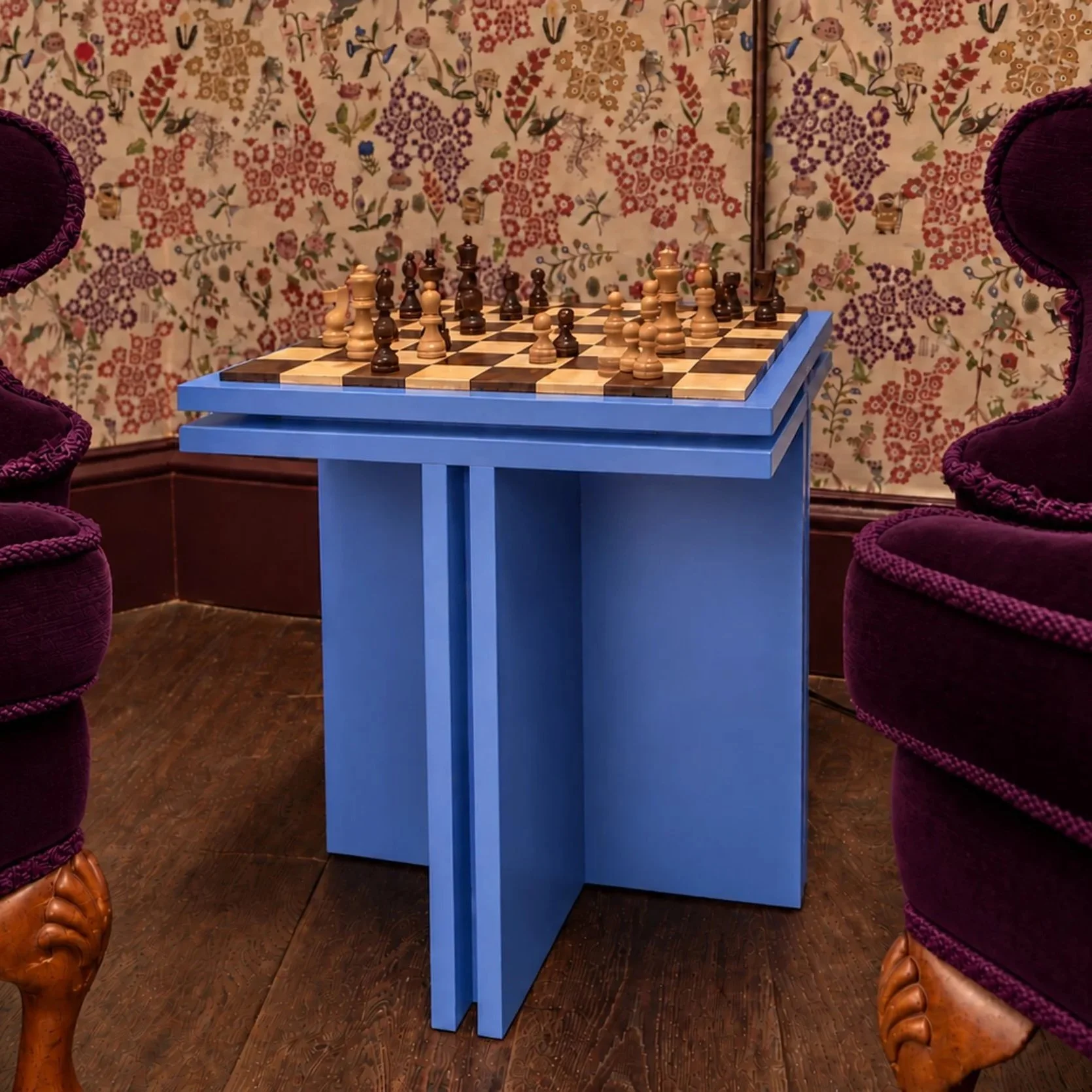 A chess table with a chessboard and pieces, surrounded by burgundy velvet chairs with carved wooden legs, set against a floral wallpaper with bird and flower motifs.