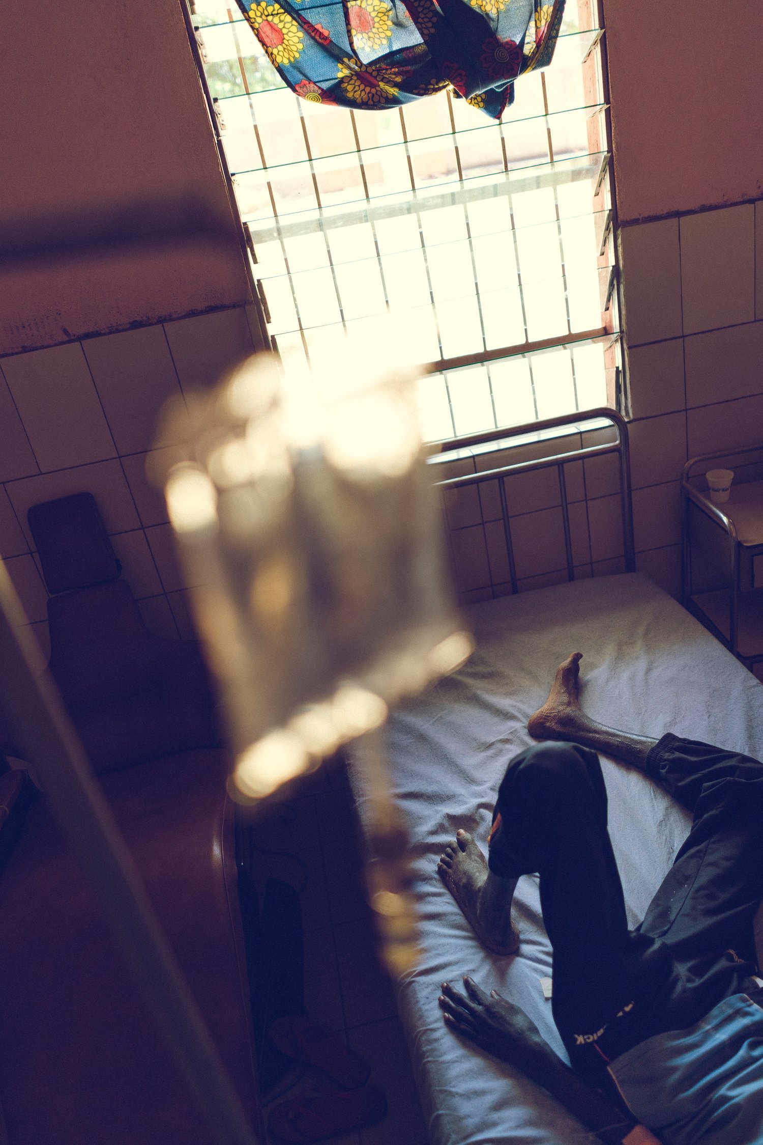 Person lying on their back in an isolated, sunlit room with open windows and bars outside; view from above evokes vulnerability and confinement, suggesting emotional or psychological distress