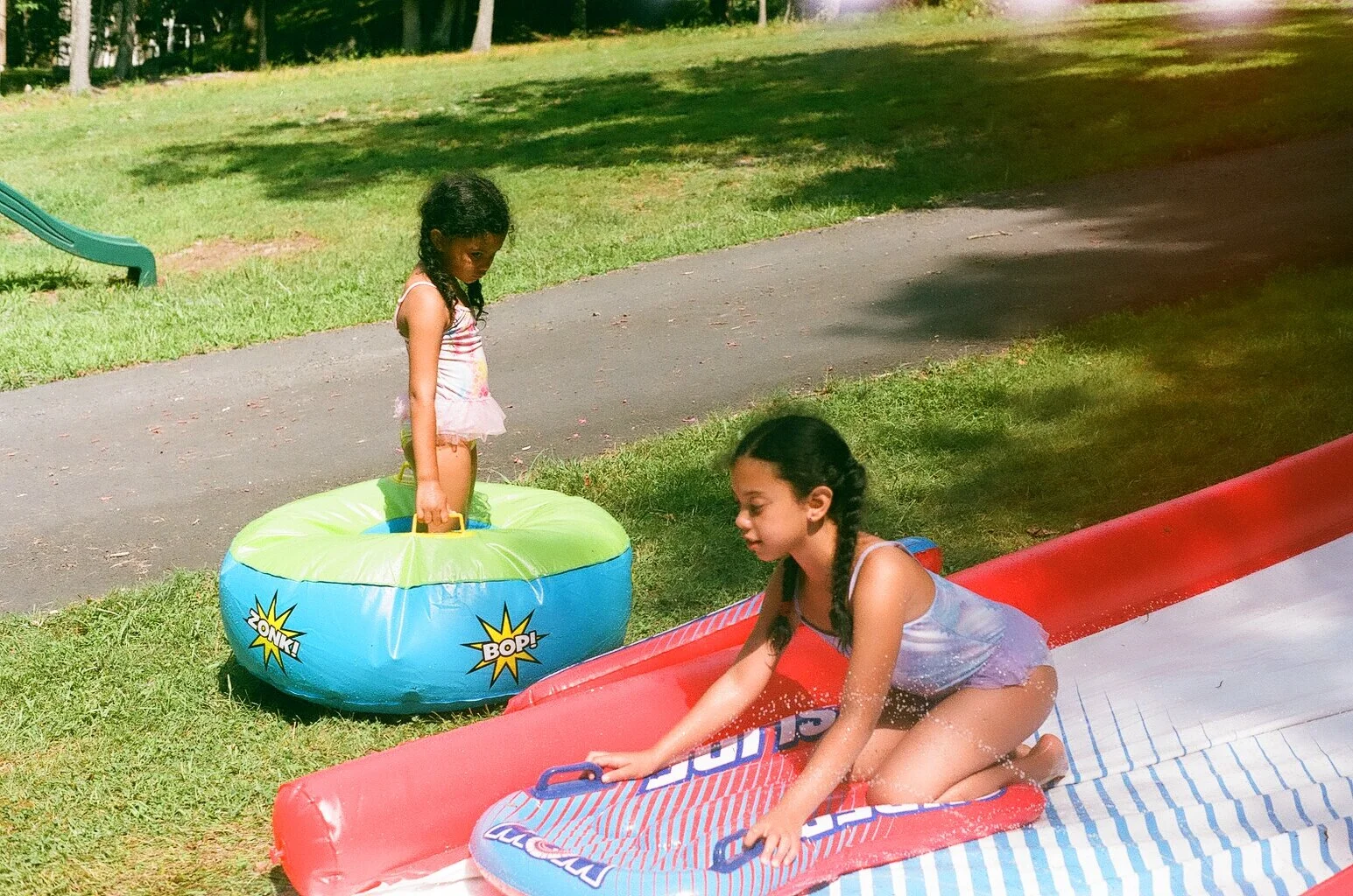 Two young girls playing on a slip and slide water slide outdoors on a sunny day, with one girl sitting on the slide and the other girl standing nearby in a splash pool.
