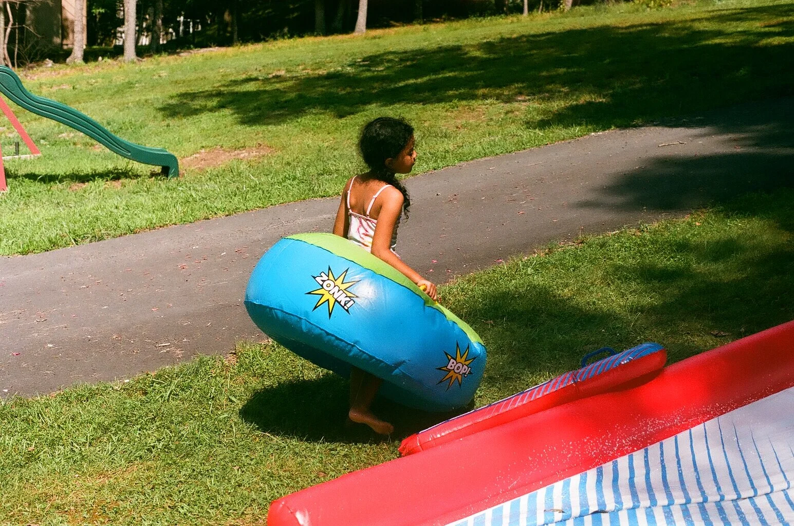 A young girl with dark hair and a striped swimsuit, holding a colorful inner tube, preparing to go down a water slide outdoors on a grassy area.