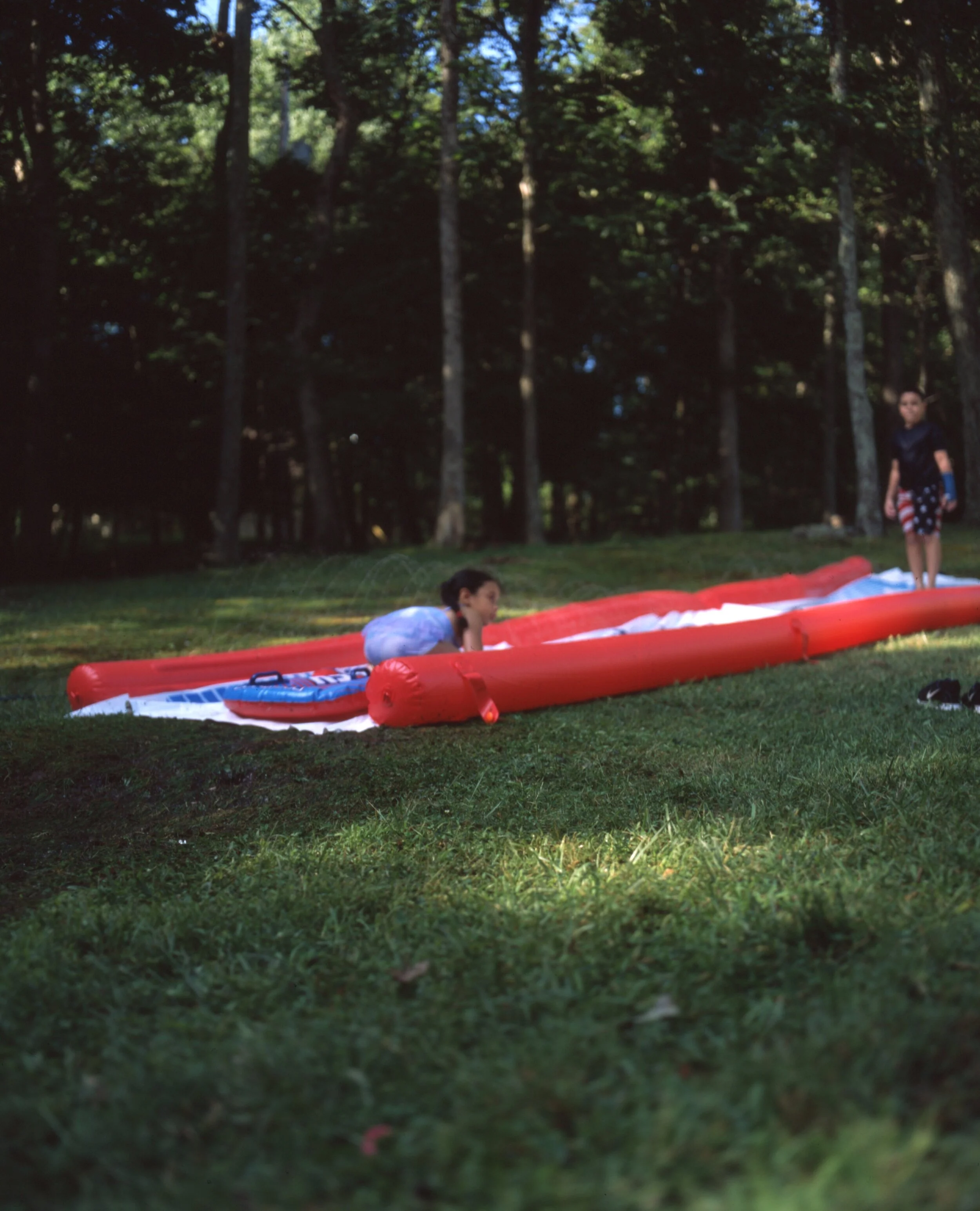 Two children playing on an inflatable slip and slide in a grassy backyard with tall trees in the background.