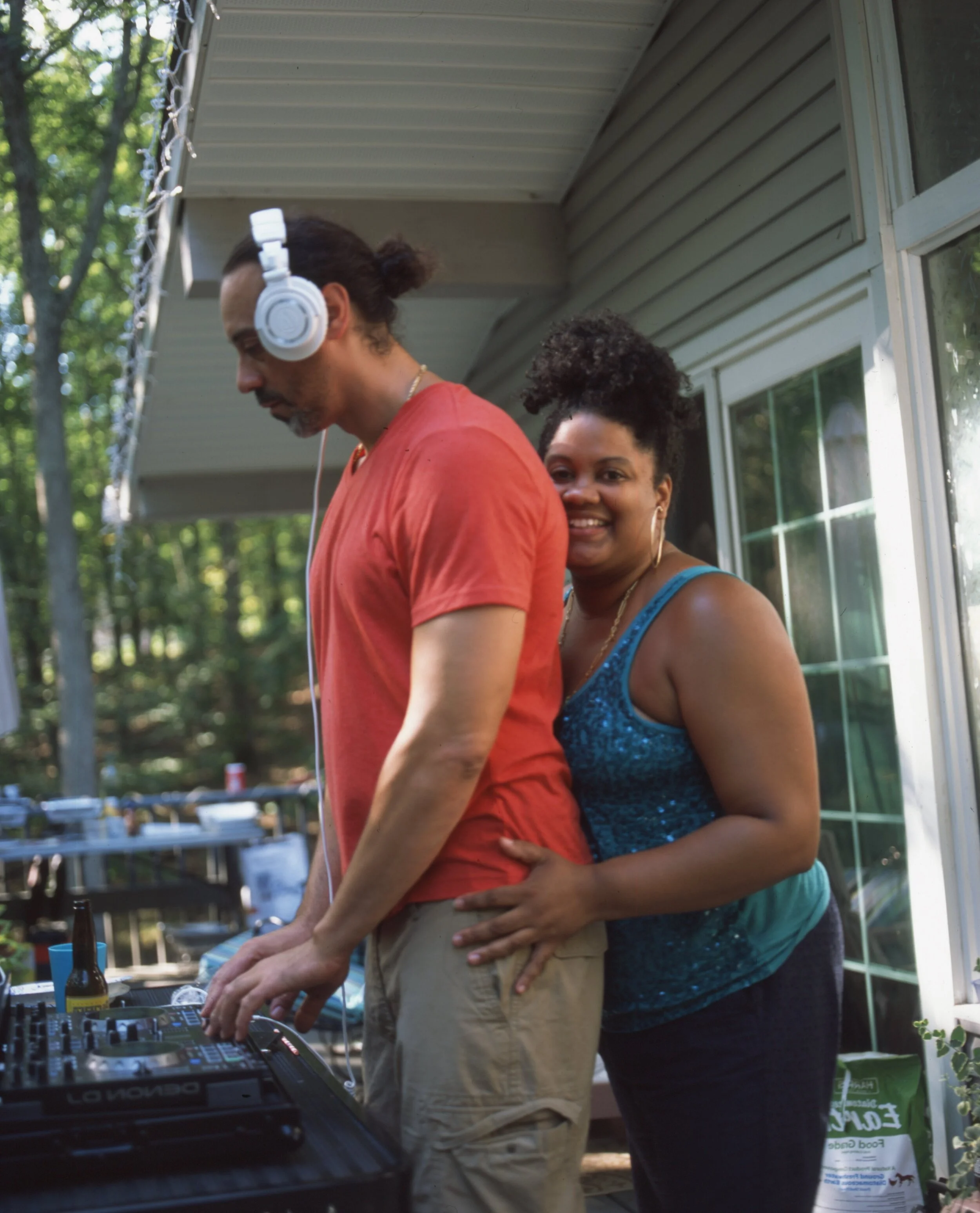A man wearing headphones and a red t-shirt standing by a DJ mixer, with a woman in a blue sparkly top standing behind him, smiling and holding his waist, outside on a porch surrounded by trees.
