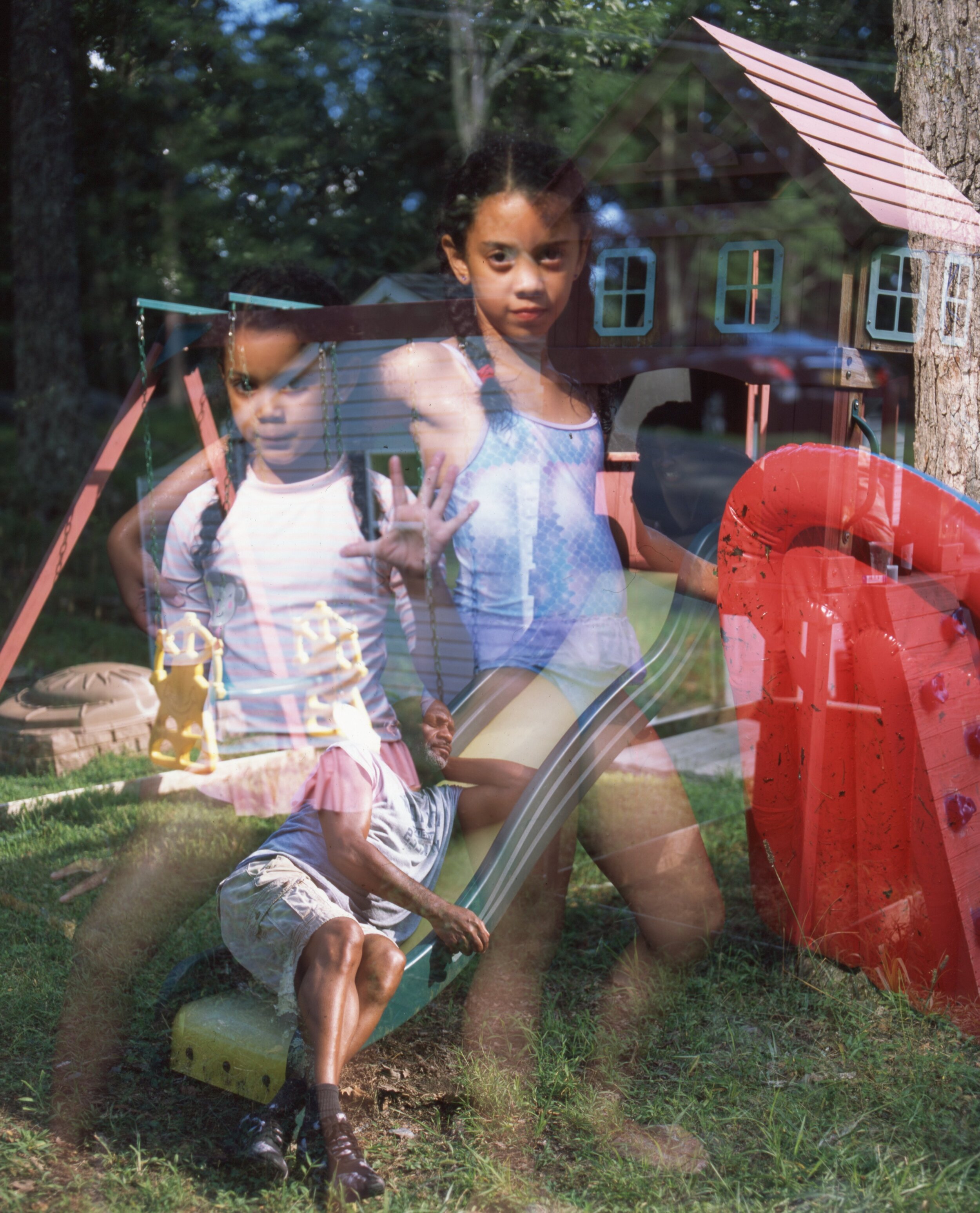 A double exposure photo of two young girls playing outside on a swing set, with a man climbing a slide below them. The background shows trees and a child's playhouse.