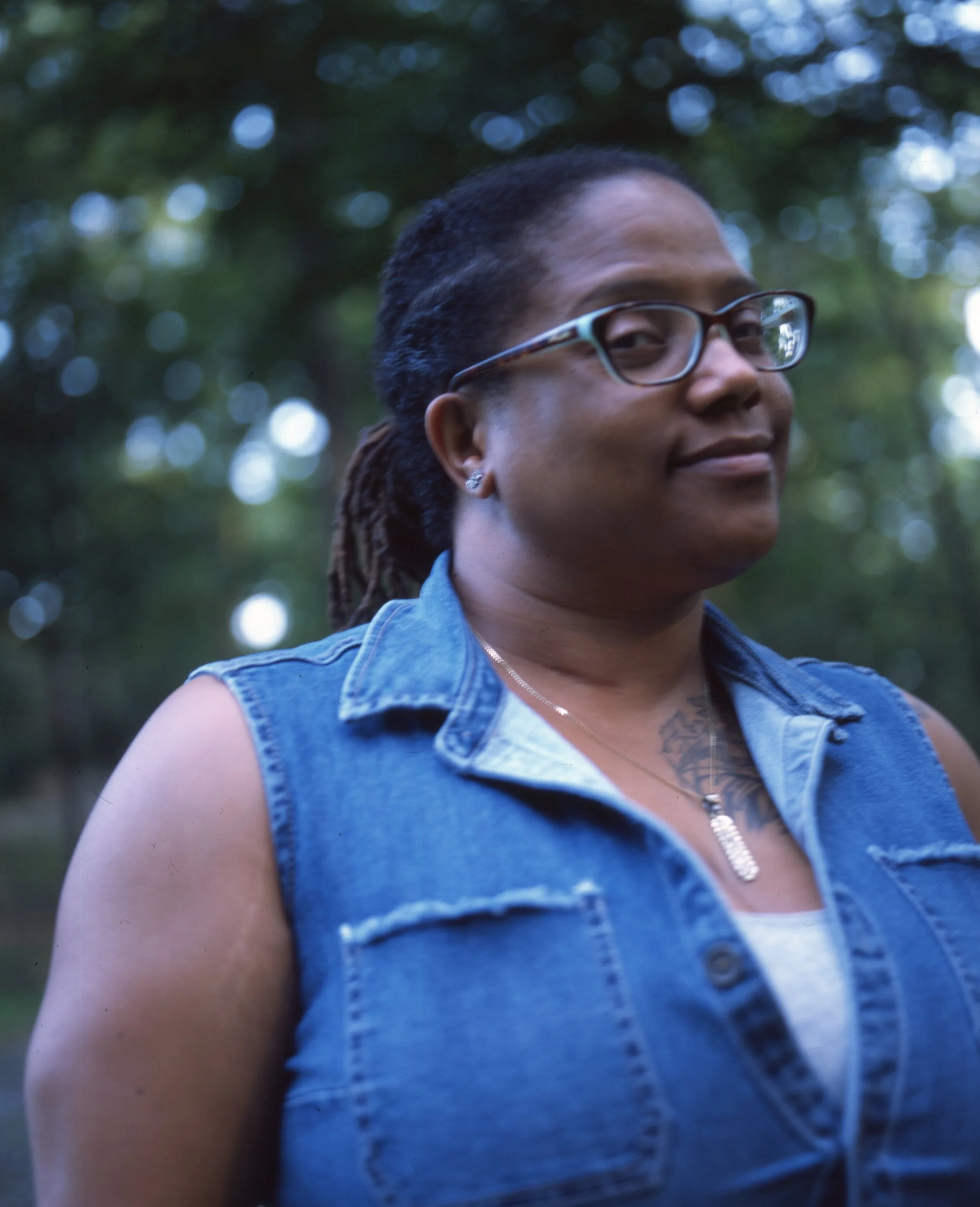 A woman with glasses and dark skin, wearing a sleeveless denim jacket, stands outdoors with a blurred background of trees and dappled sunlight.