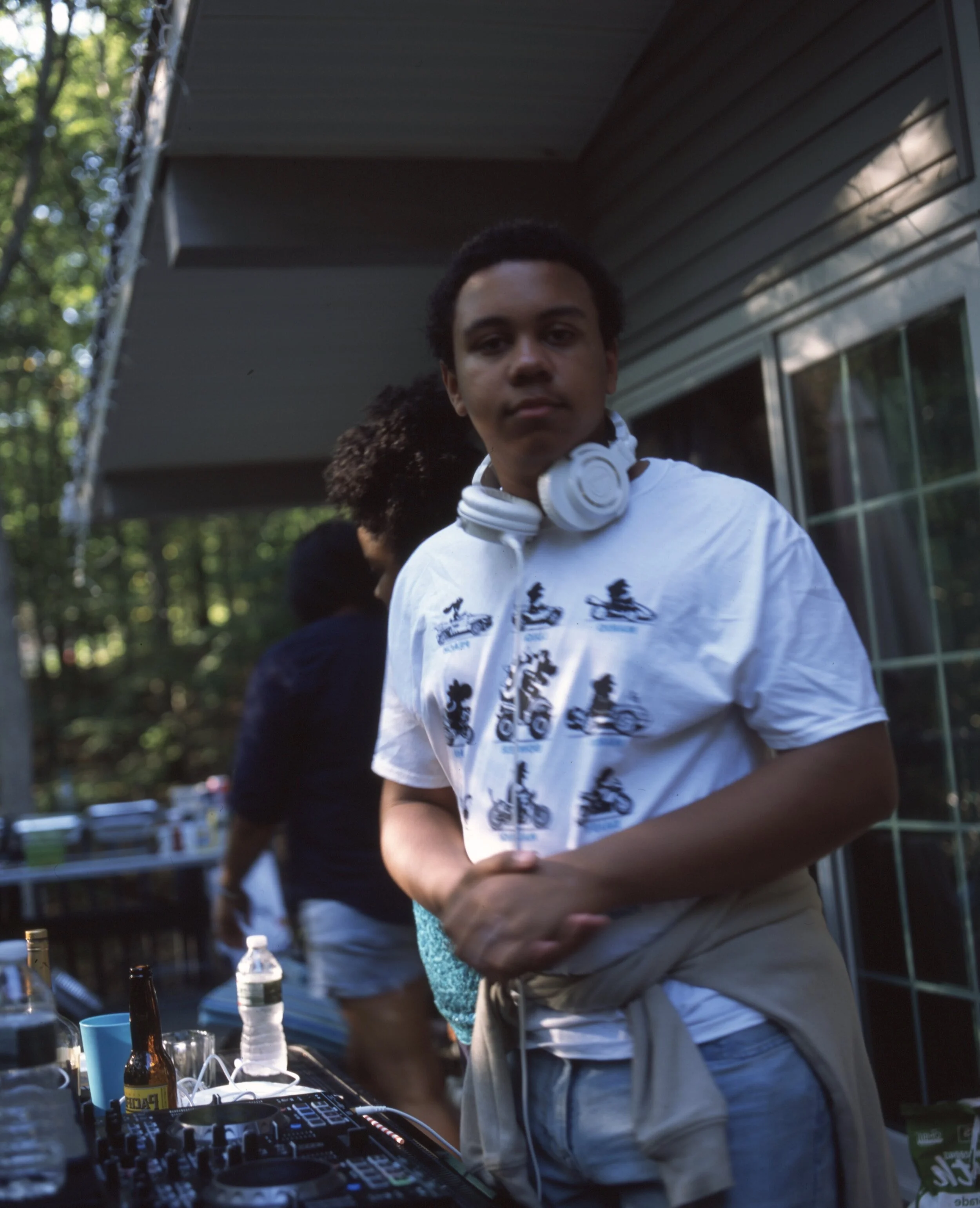 A young man wearing a white DJ shirt with headphones around his neck stands outside near a table with drinks and DJ equipment.