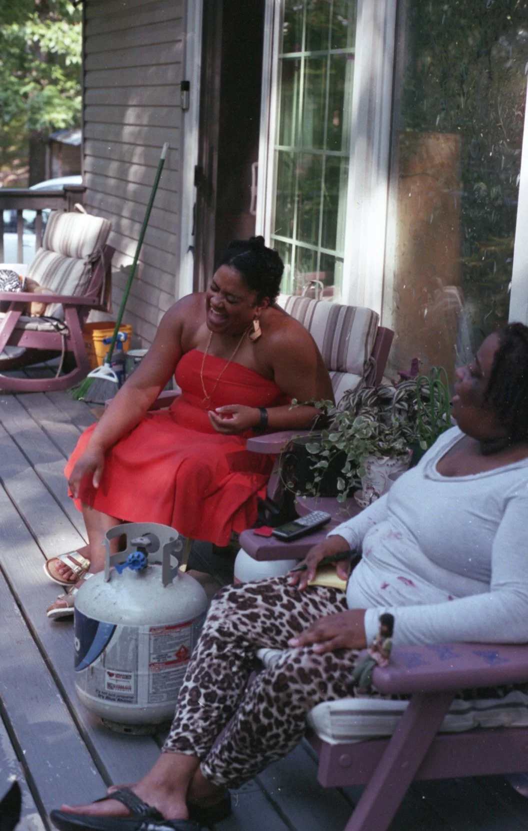 Two women sitting outside on a wooden deck, engaged in conversation and laughter. One woman is in a red strapless dress, and the other is in a white long-sleeve shirt and animal print pants. Items around them include a propane tank, a remote control,