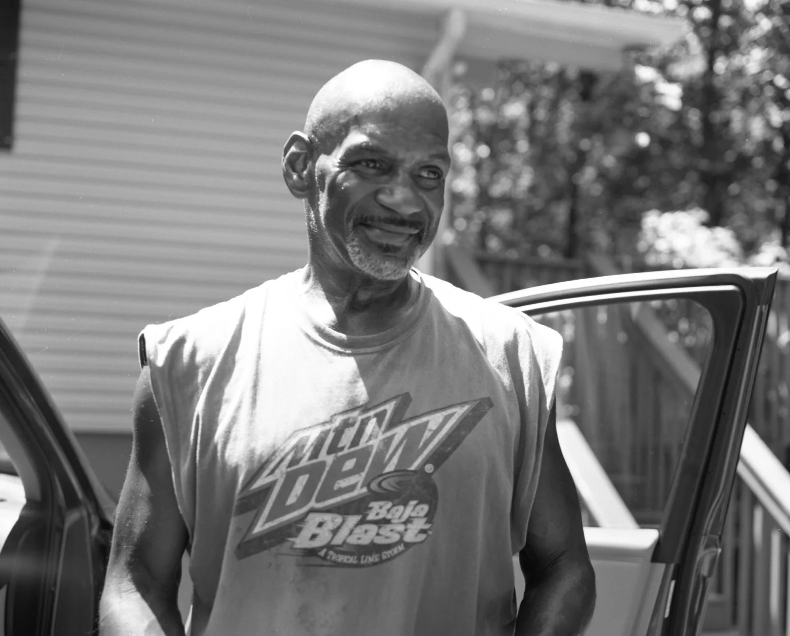 An elderly man with a bald head, gray beard, and smiling face standing outside next to a car door, wearing a sleeveless T-shirt with a logo on it.