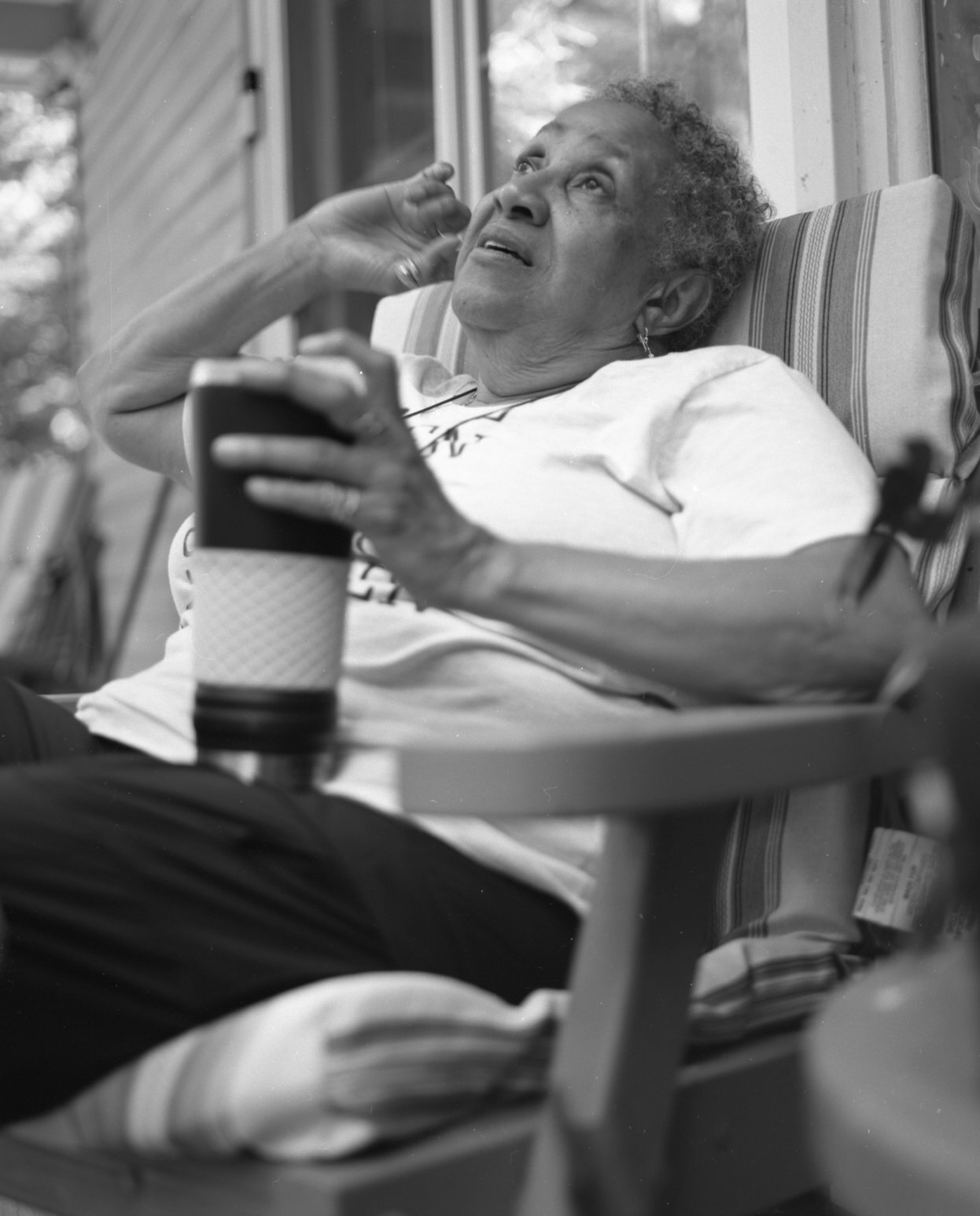 An elderly woman with short curly hair lying on a chair, holding a phone in one hand and a cup in the other, looking upwards with a concerned expression.