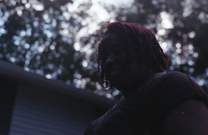 A woman with curly hair outdoors at dusk, with trees in the background.