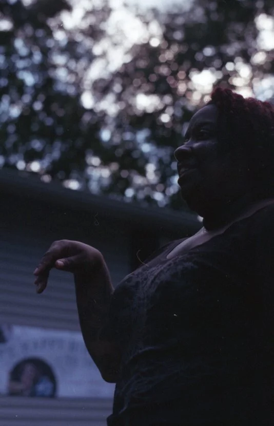 A woman standing outdoors with trees and a blurred building in the background, her face in profile and her hand raised slightly.