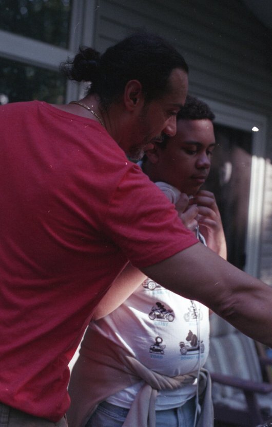 Two people, a man with a bun hairstyle and a woman, are looking at something outside the frame. The man is wearing a red shirt and the woman is wearing a white shirt with motorcycle prints, standing close together during what appears to be dusk or ev