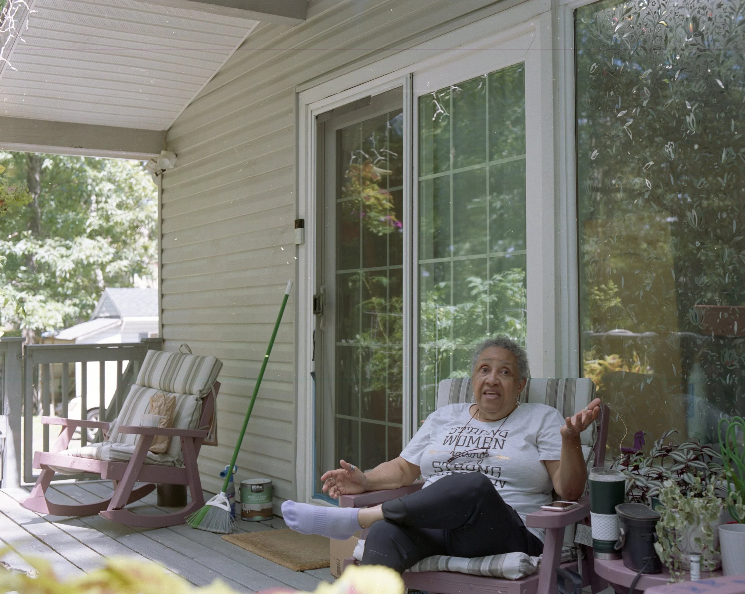 An elderly woman sitting on a porch chair, gesturing with her right hand while talking, surrounded by plants and cleaning supplies, with glass doors and a rocking chair in the background.