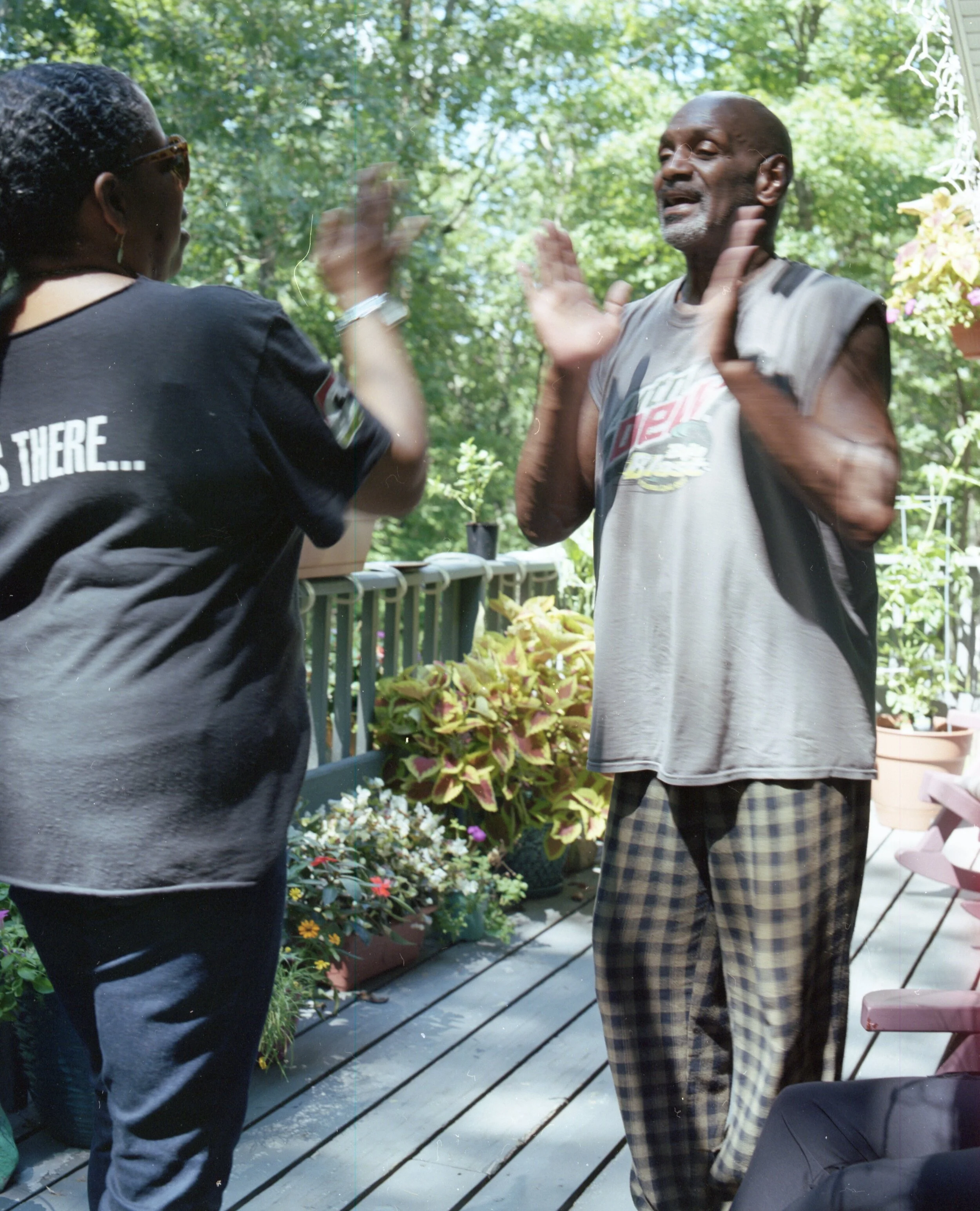 Two people high-fiving on a porch with potted plants and trees in the background.
