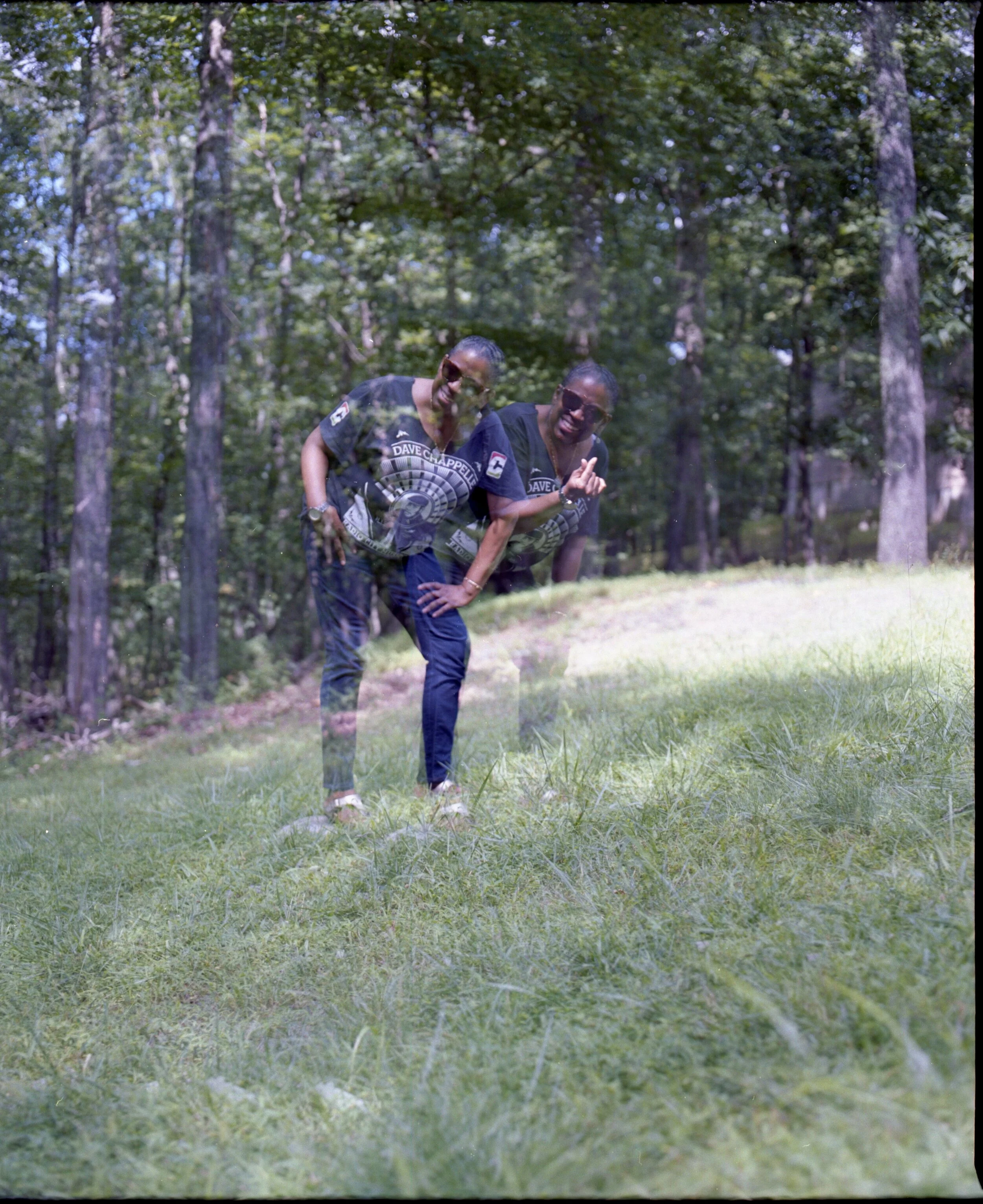 A double-exposed photo of a woman bending over and smiling, wearing sunglasses and a black T-shirt with a circular logo, outdoors in a wooded area with green grass and trees.