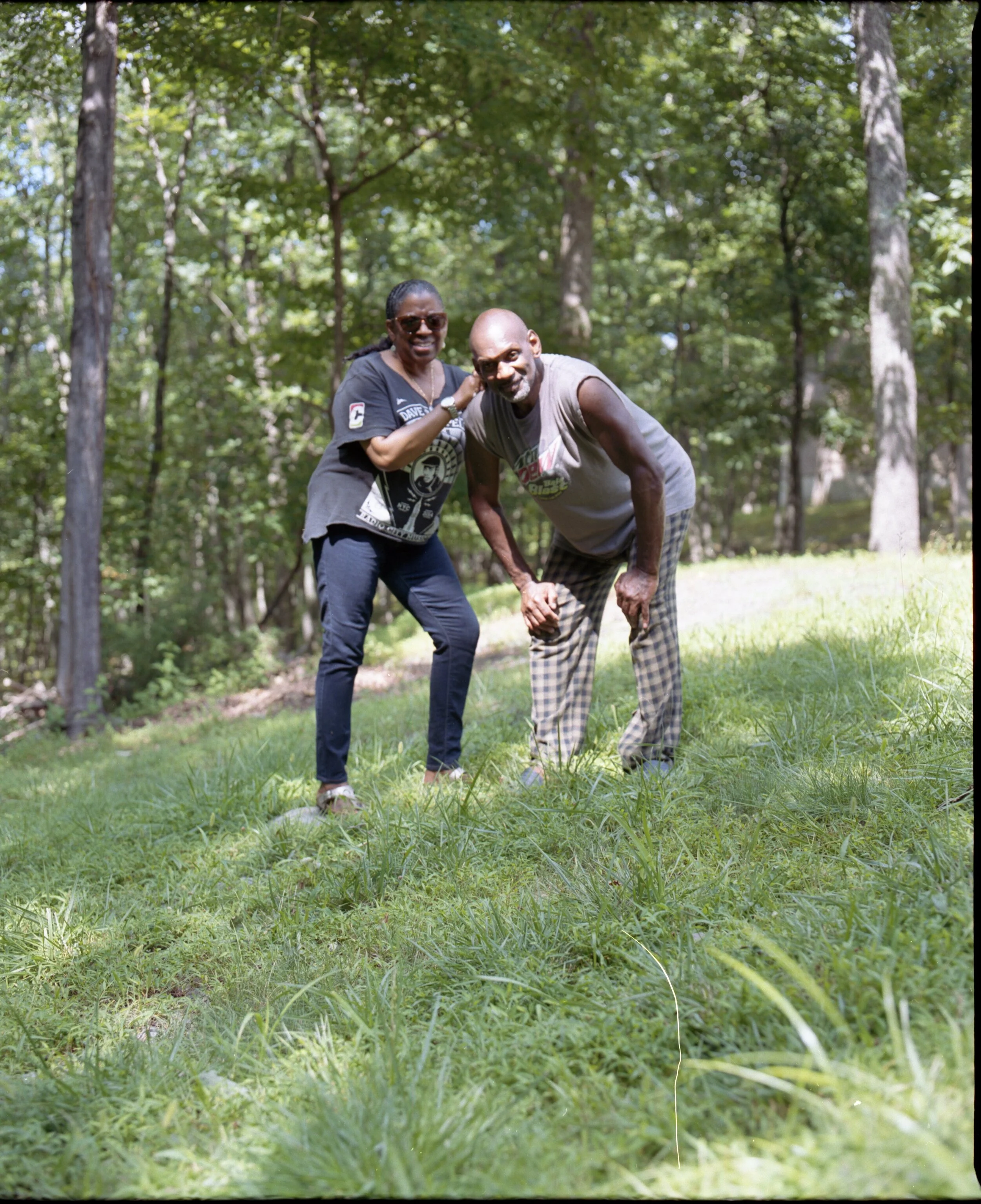 Two smiling people, a woman and a man, standing outdoors in a wooded area, with the woman shoulder-bumping the man, both leaning forward with the man bending his knees slightly and the woman placing her hand on his shoulder.