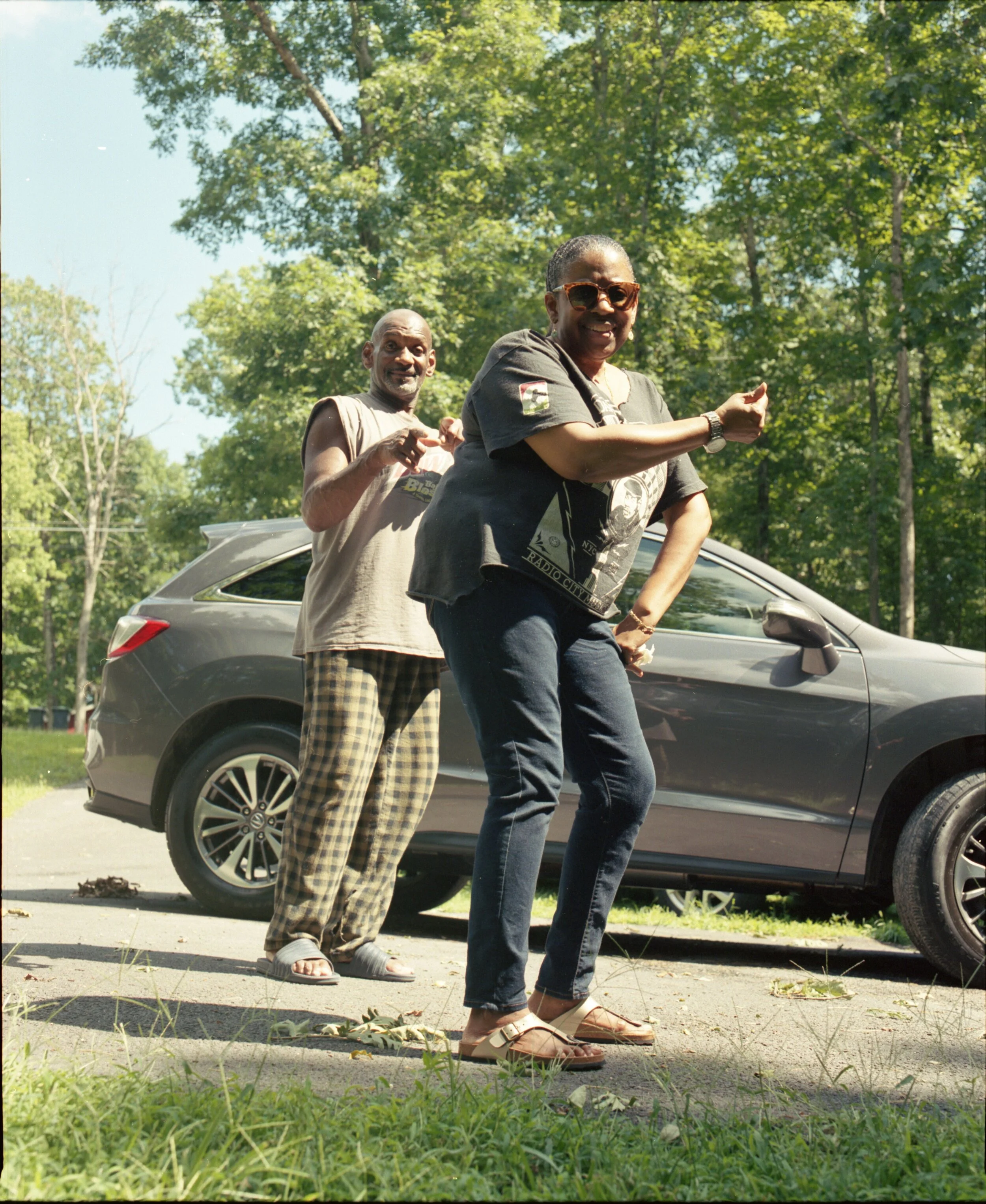 Two people dancing outdoors near a gray car, with a man behind a woman, both smiling. The woman wears sunglasses and casual clothing, and the man wears a sleeveless shirt and plaid pants. There are trees and grass in the background.