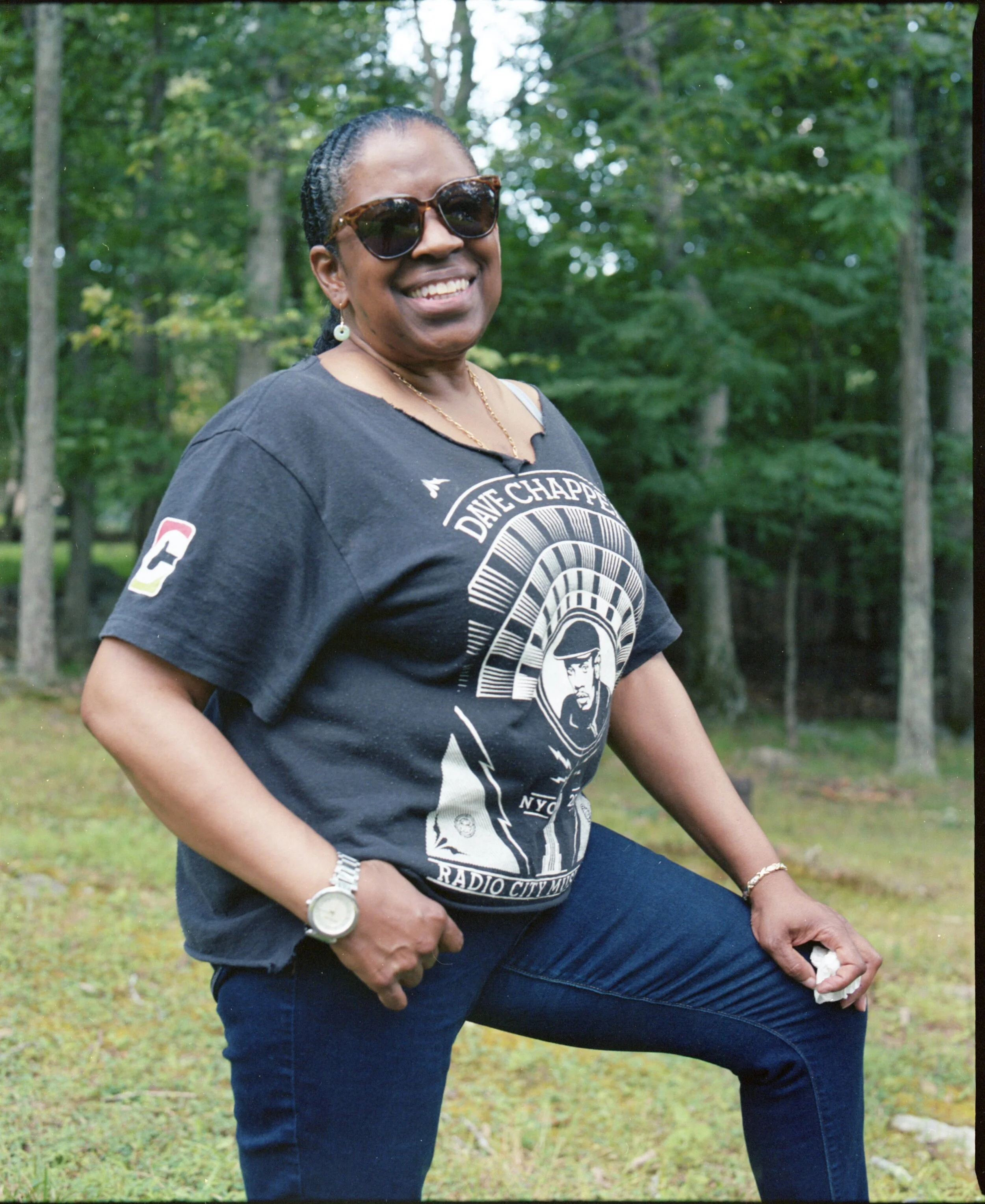 A smiling woman with dark sunglasses, wearing a dark t-shirt featuring artwork of a man and text, standing outdoors in front of trees.