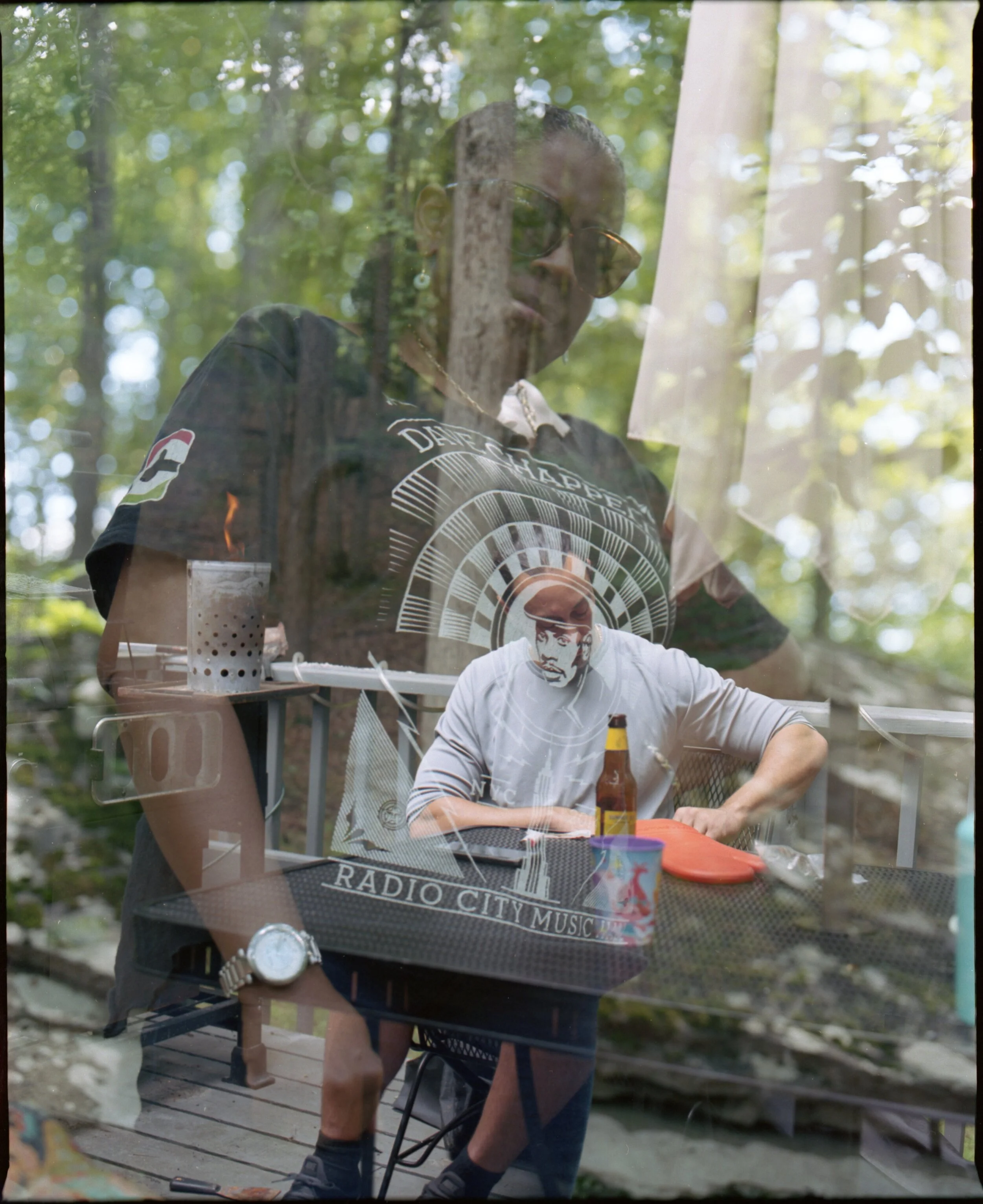A woman with sunglasses and earrings seen through a window, with her reflection overlapping a man sitting outdoors, who is wearing a white shirt, black shorts, and a paper mask, sitting at a table with a beer bottle and a cup.