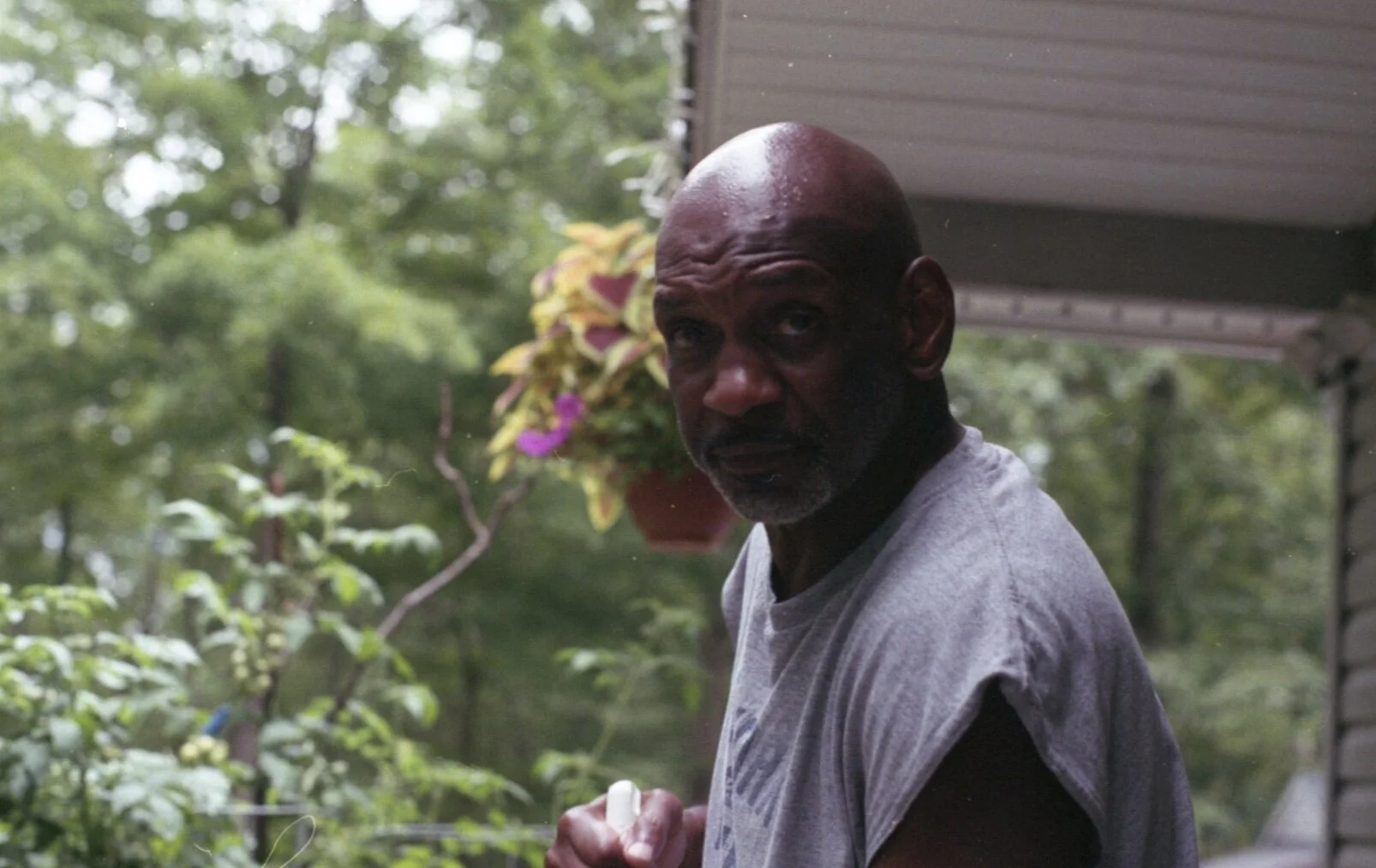 A bald man with a serious expression, wearing a gray sleeveless shirt, stands outdoors near a house with a porch and trees in the background.