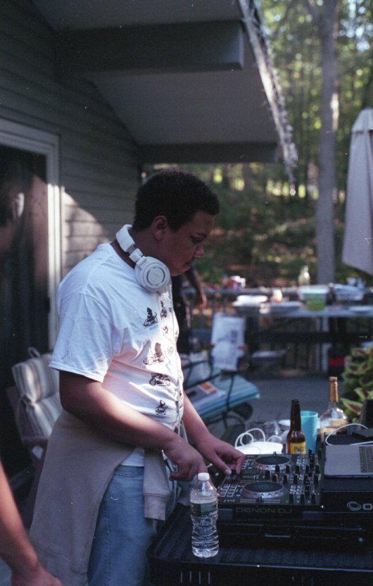 A young male DJ wearing a white t-shirt with headphones around his neck, operating DJ equipment outdoors on a deck during the daytime.
