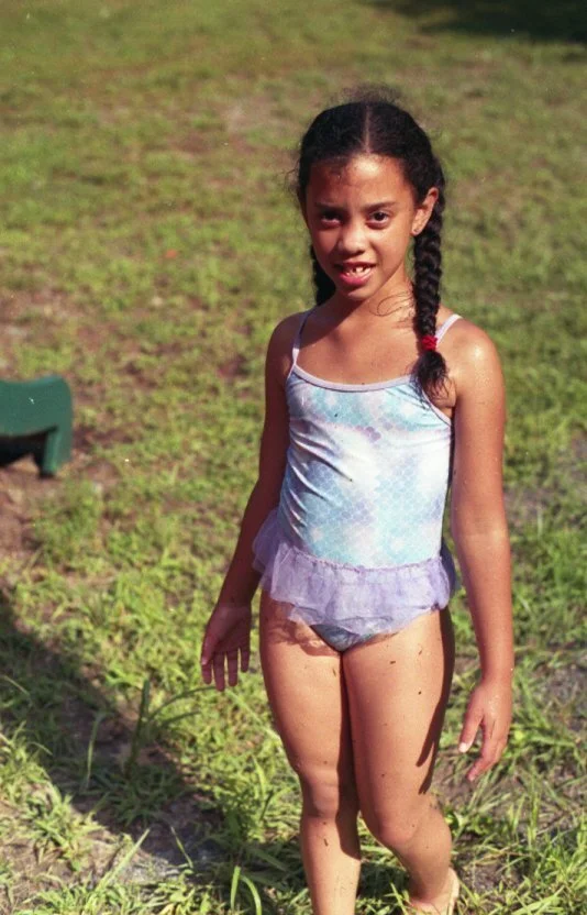Young girl with dark hair in braids outside on grass, wearing a light blue sleeveless top with ruffled hem and matching bottoms