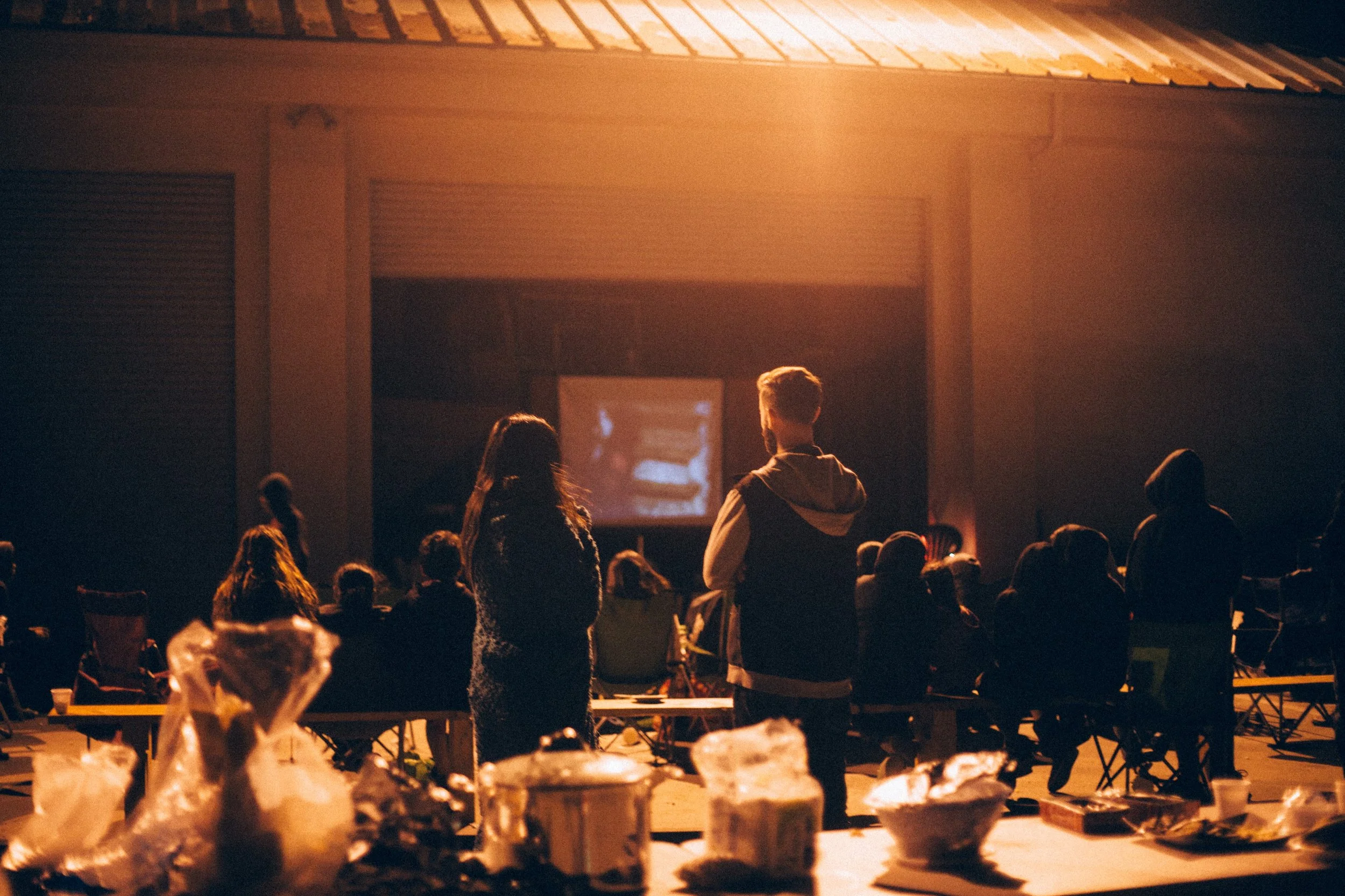 People watching a film at an outdoor theater during the evening, with snacks and drinks on tables in the foreground.