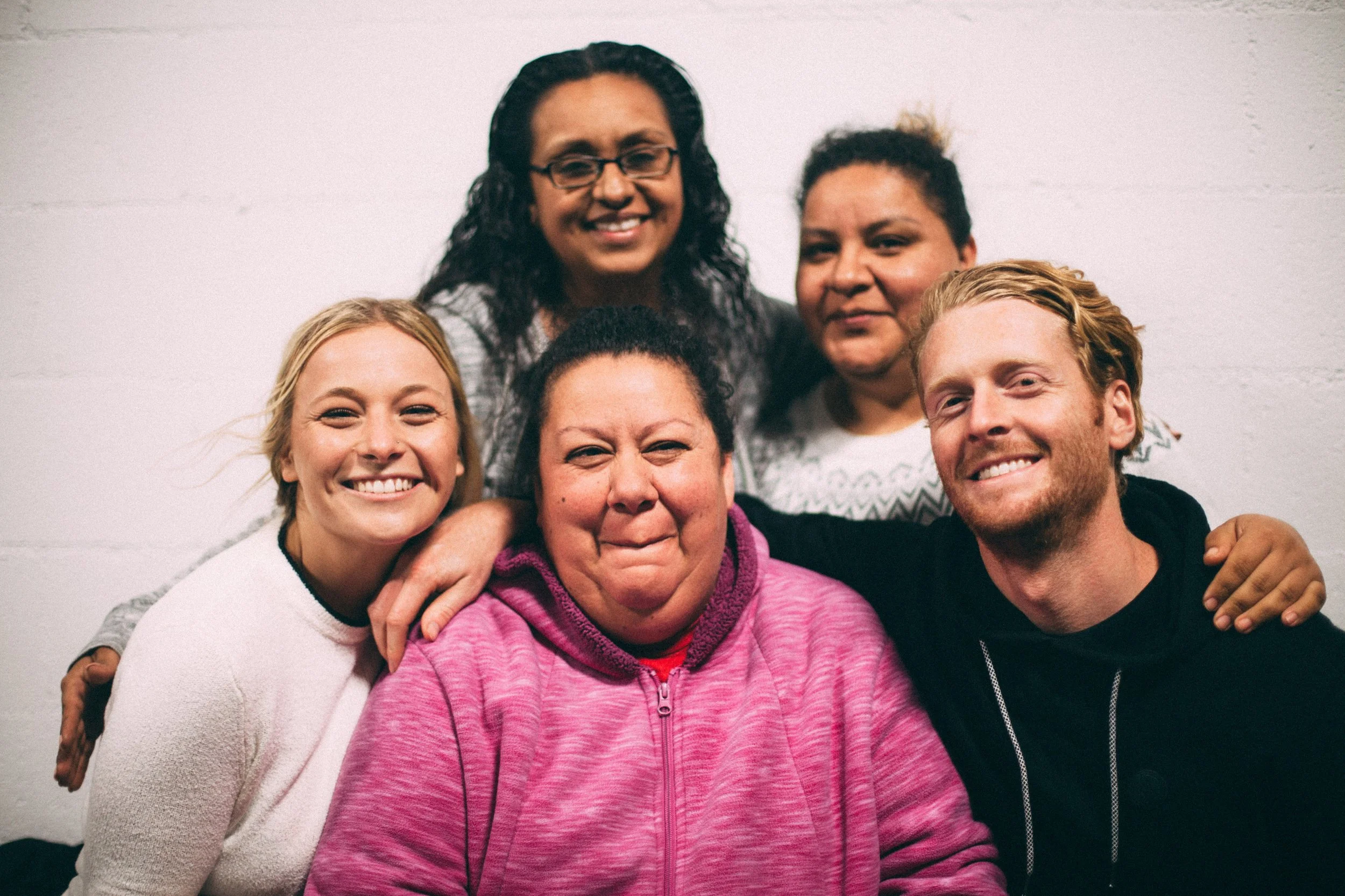 Group photo of six diverse smiling people in casual clothing, standing close together against a plain white wall.
