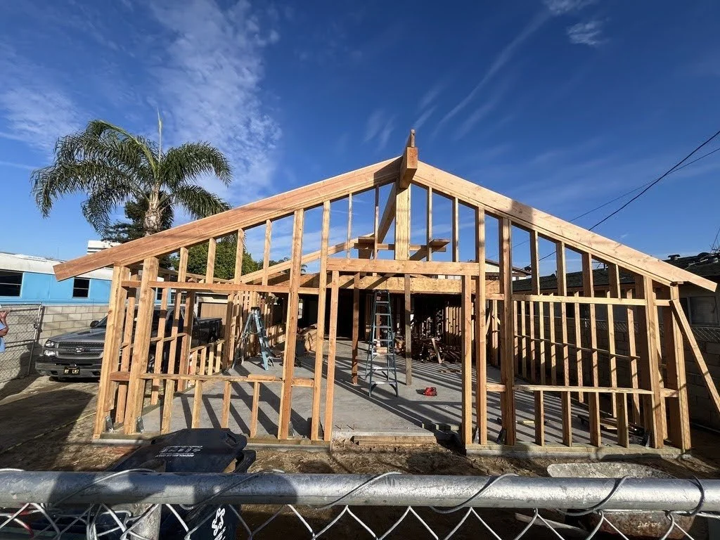 Construction site with wooden framing for a house, clear blue sky with some clouds, a tree, parked cars, and construction tools visible.