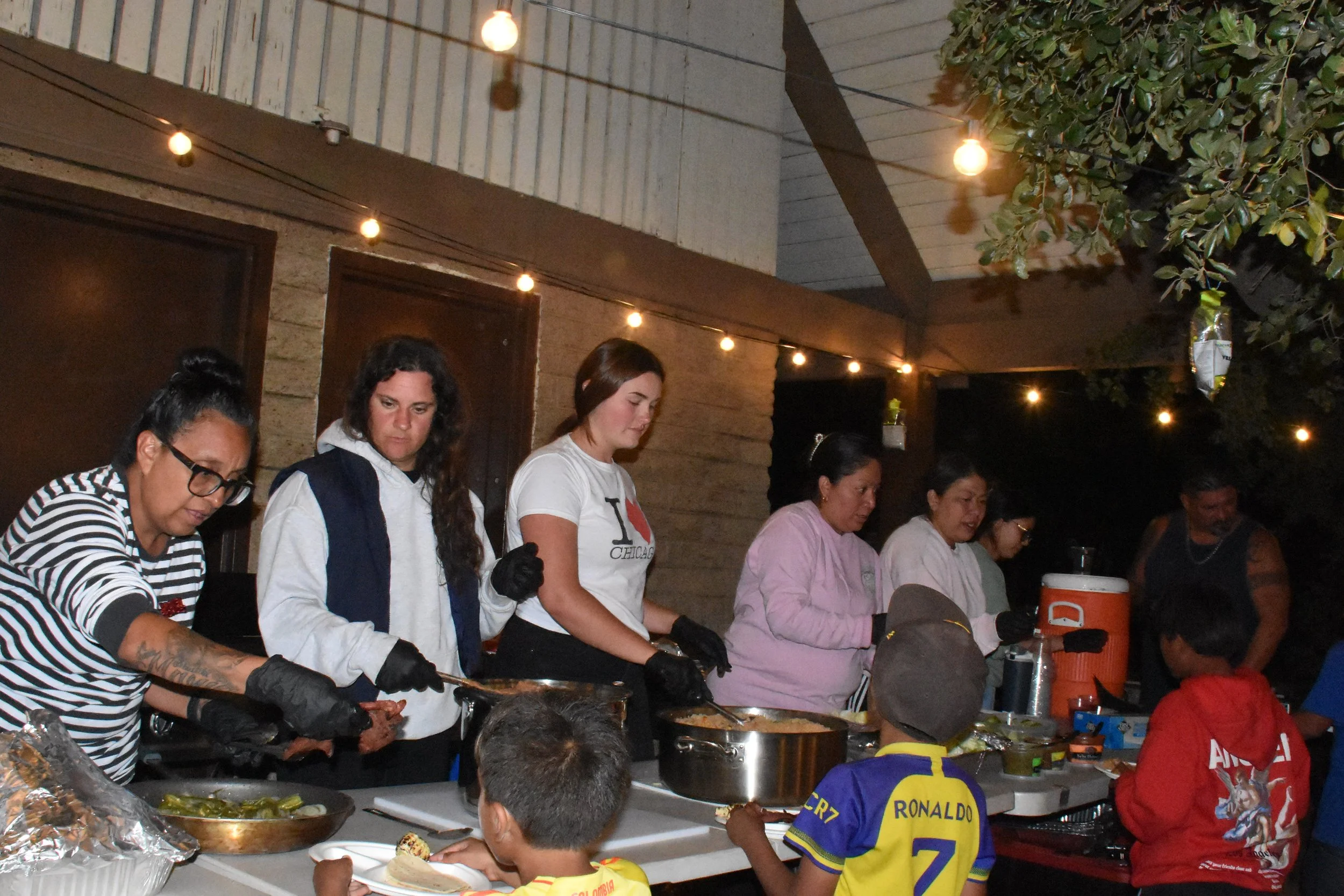 An outdoor evening event with numerous people sitting at tables with lit candles, string lights above, in an urban setting with modern buildings and a large LED screen.