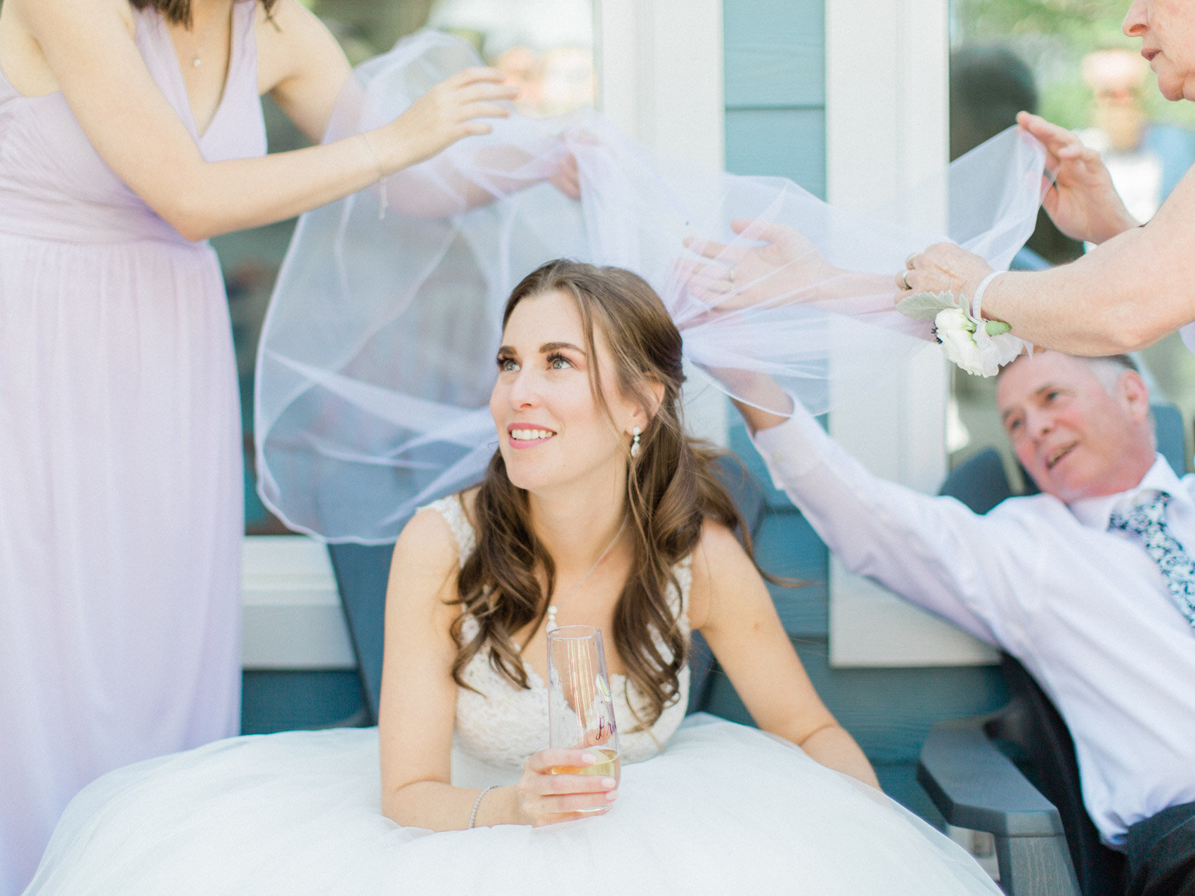 relaxed bride enjoying a glass of champagne while friends and family pick bugs out of her veil at her summer muskoka cottage wedding