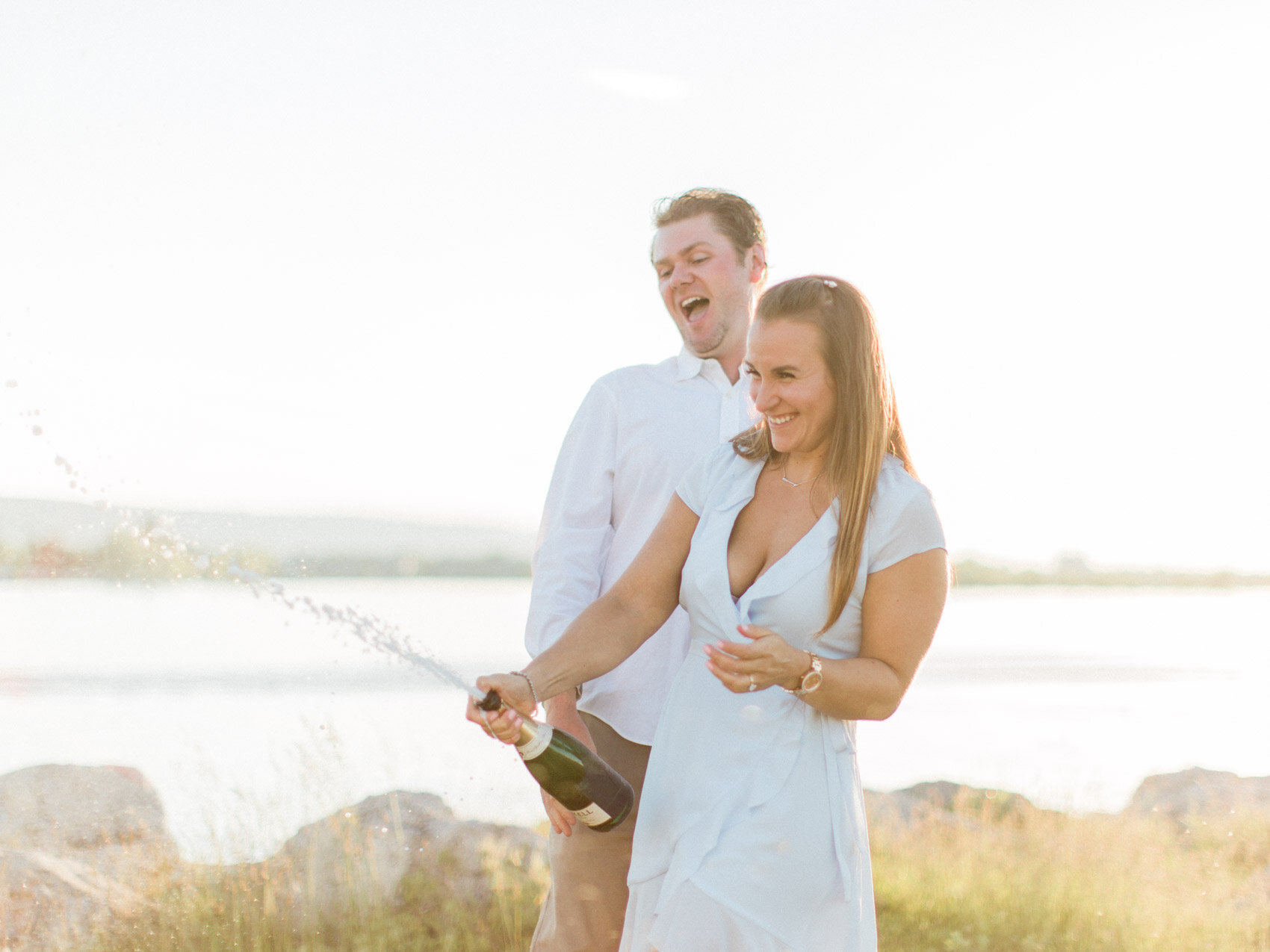 cute excited couple popping a bottle of champagne after he proposed at the collingwood waterfront at sunset