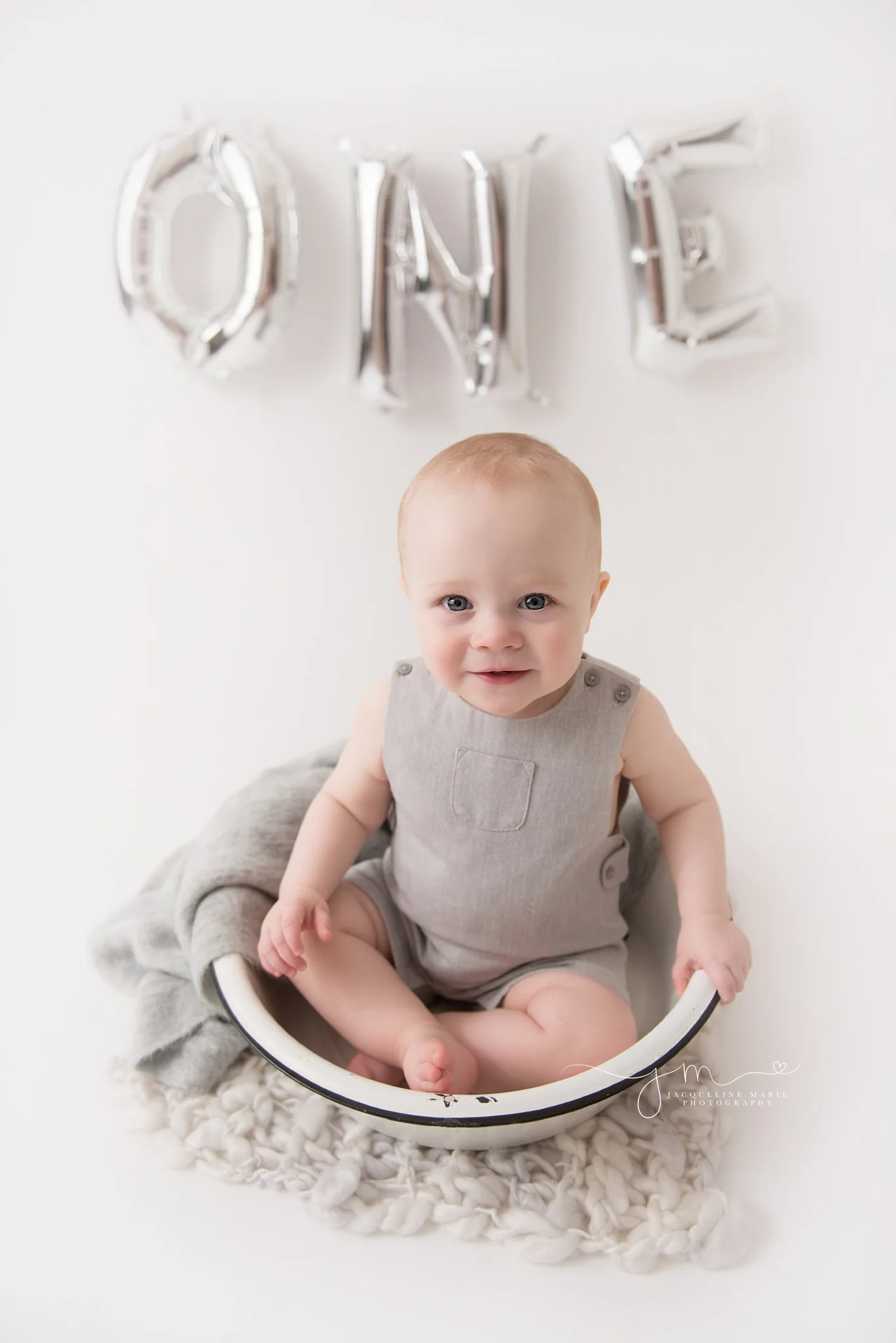 one year old baby boy wears gray romper while sitting in antique bowl for first birthday portraits in columbus ohio