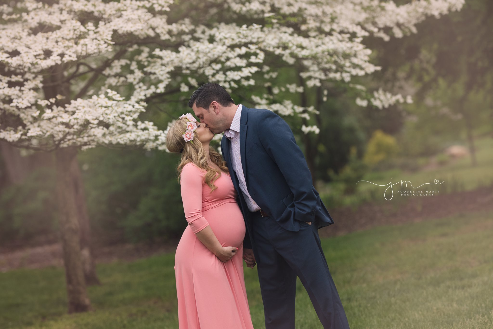 expecting parents share a kiss in park with white floral tree while wearing navy and coral in columbus ohio for maternity pictures