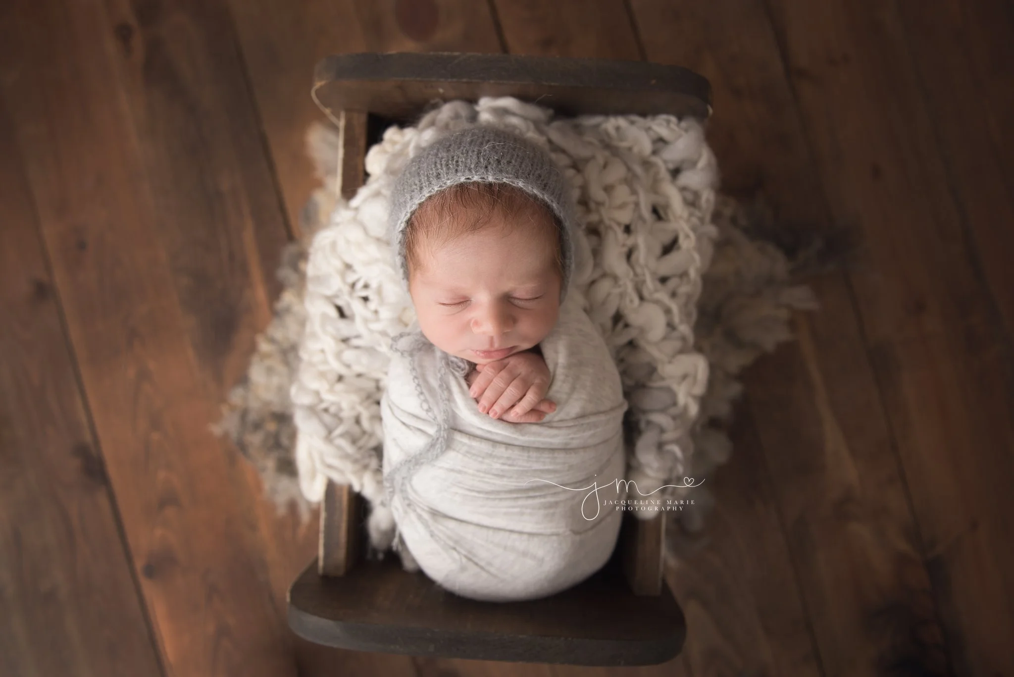 newborn baby boy wears gray bonnet while swaddled in wooden bed for newborn photography session in columbus ohio
