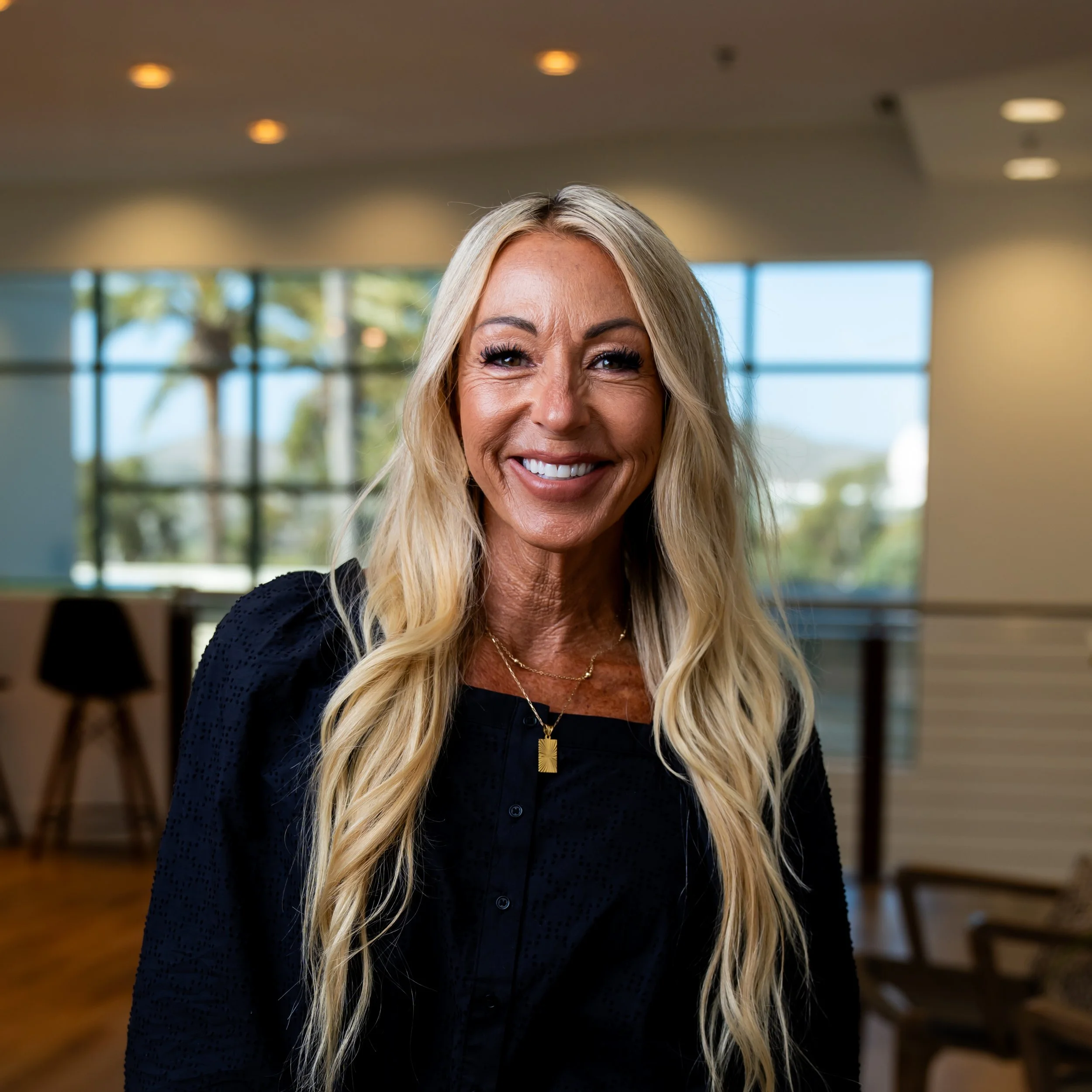 A smiling woman with long blonde hair, wearing a black top and gold jewelry, standing in a modern indoor space with large windows and outdoor trees visible in the background.
