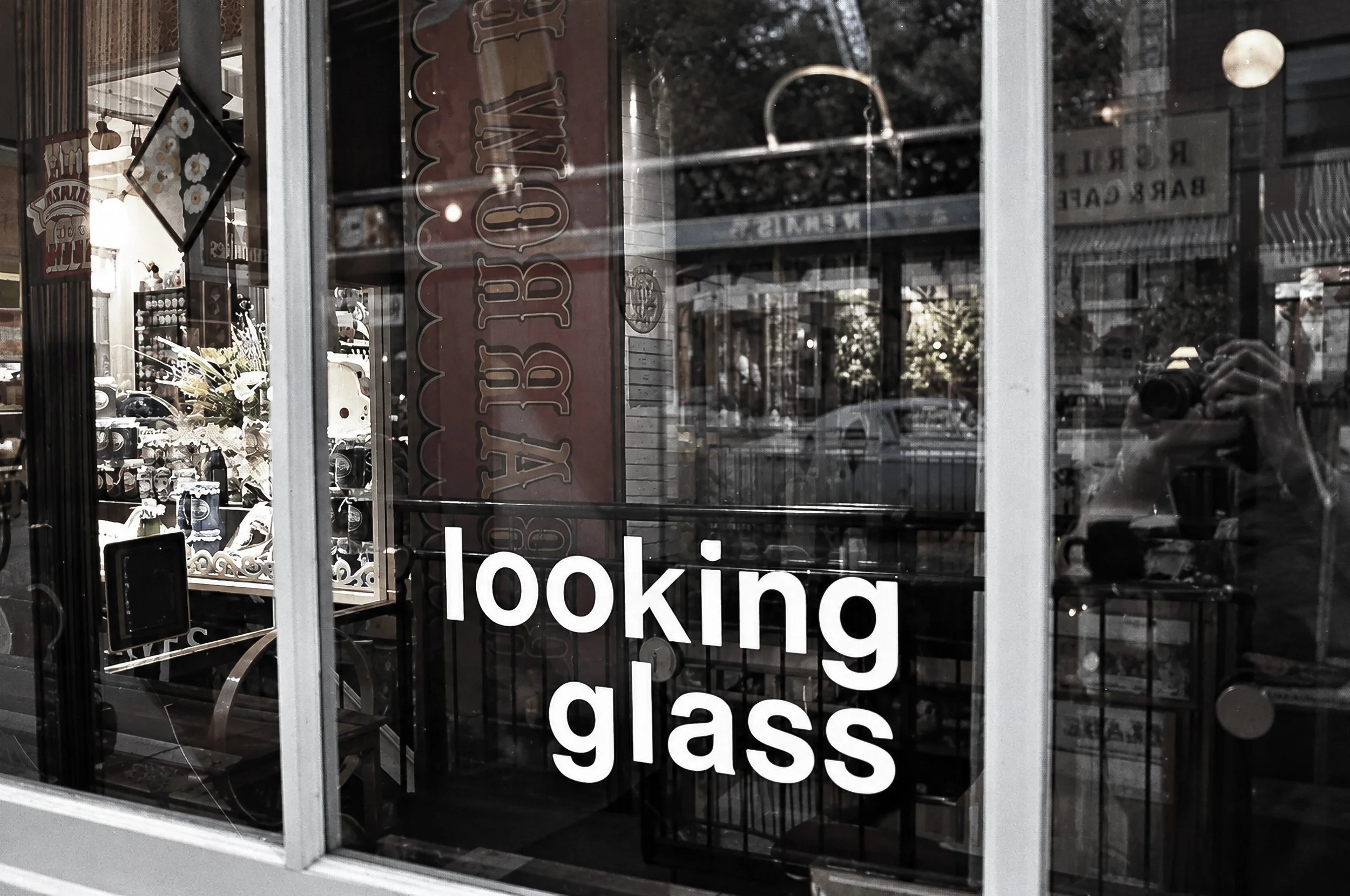 Looking Glass - The words “looking glass” appear across a shop window, layered over reflections and the objects displayed inside. Glass, lettering and reflected street life combine to create a strongly graphic photograph.