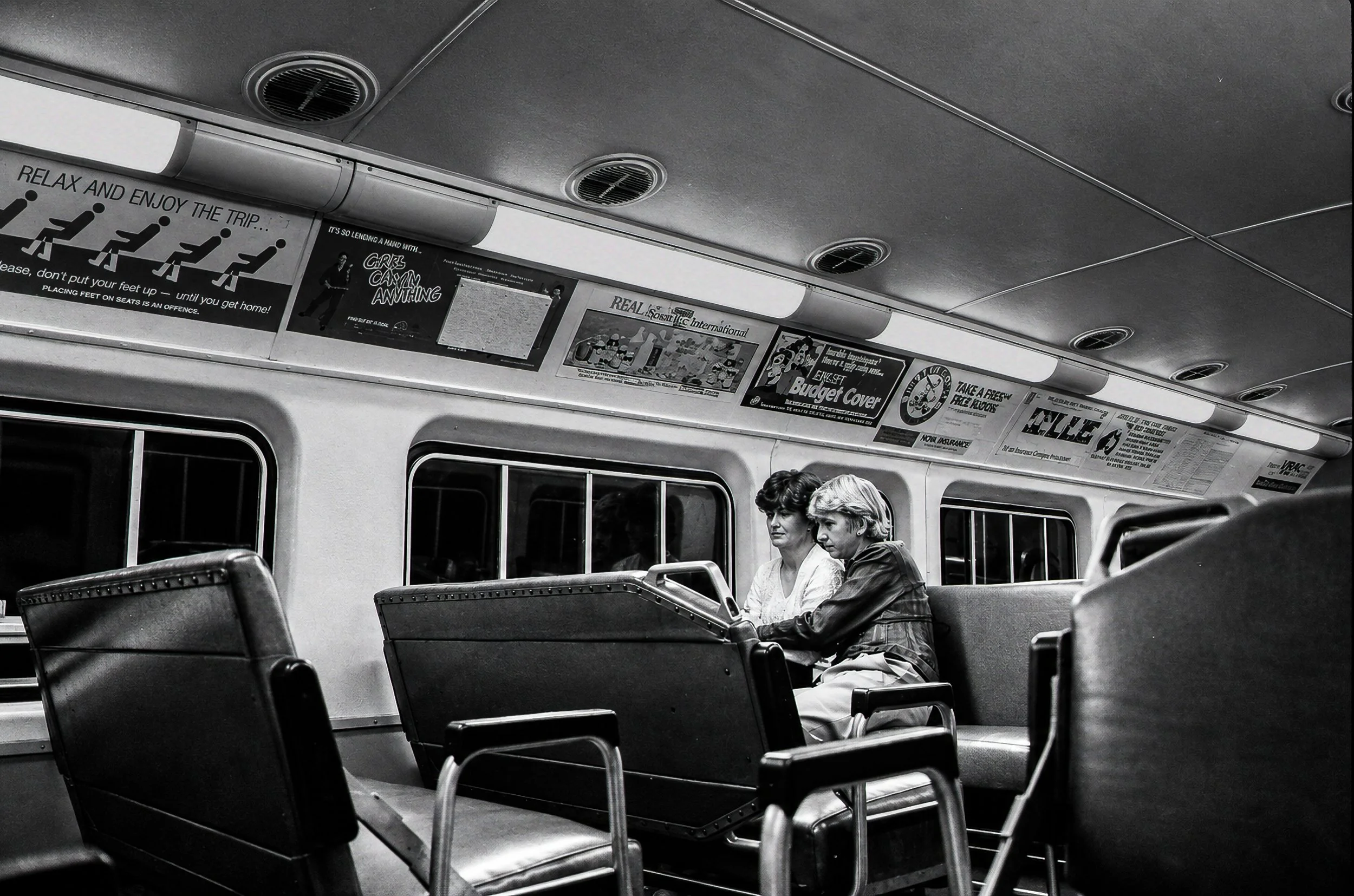 The Quiet Carriage - Two passengers sit together inside a Sydney train carriage while the empty seats and period advertisements surround them. The photograph records the understated atmosphere of public transport away from the crowds of the platform.