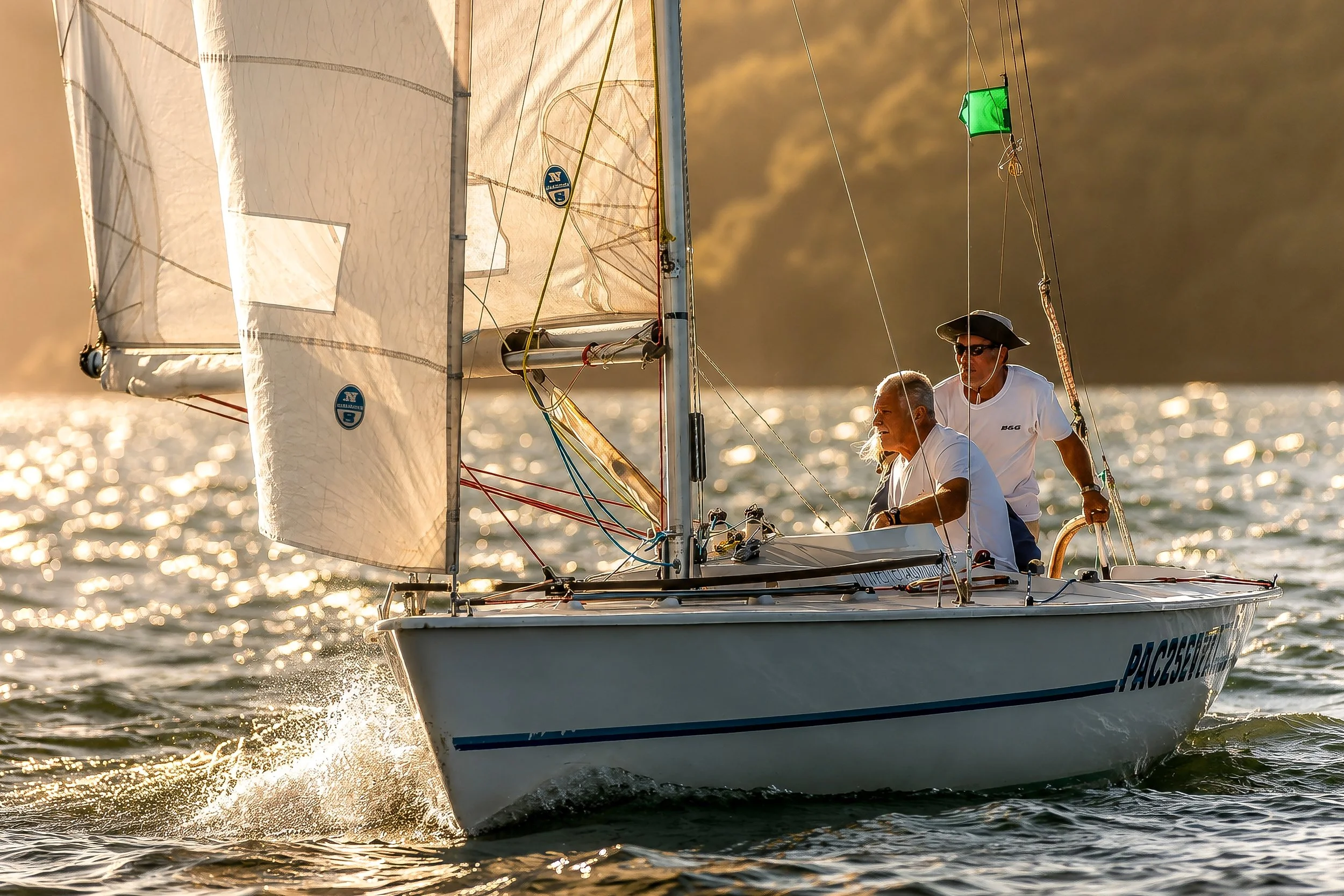 A sailboat with two people sailing on the water during sunset, with sunlight reflecting off the surface and sails catching the wind.
