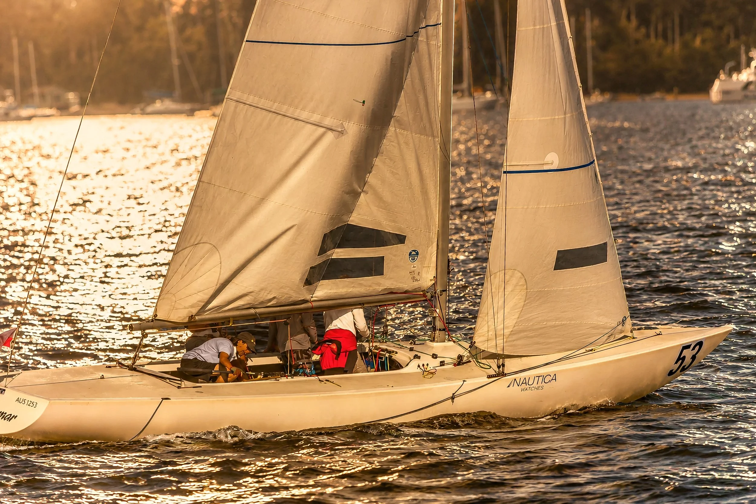 A sailboat with beige sails on a body of water during sunset, with several crew members below deck and the water reflecting the warm sunlight.