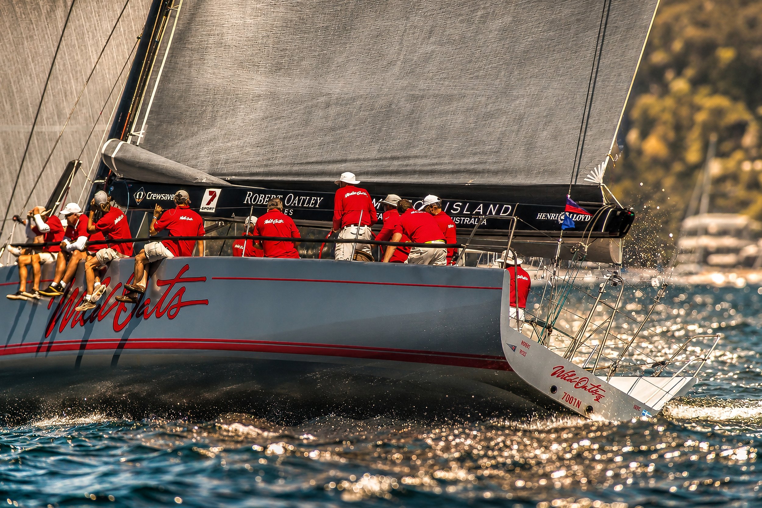 A sailing yacht with a large grey sail named 'Wild Oats XI' racing on the water, crew members in red shirts sitting on the edge of the boat, and a scenic background with trees.