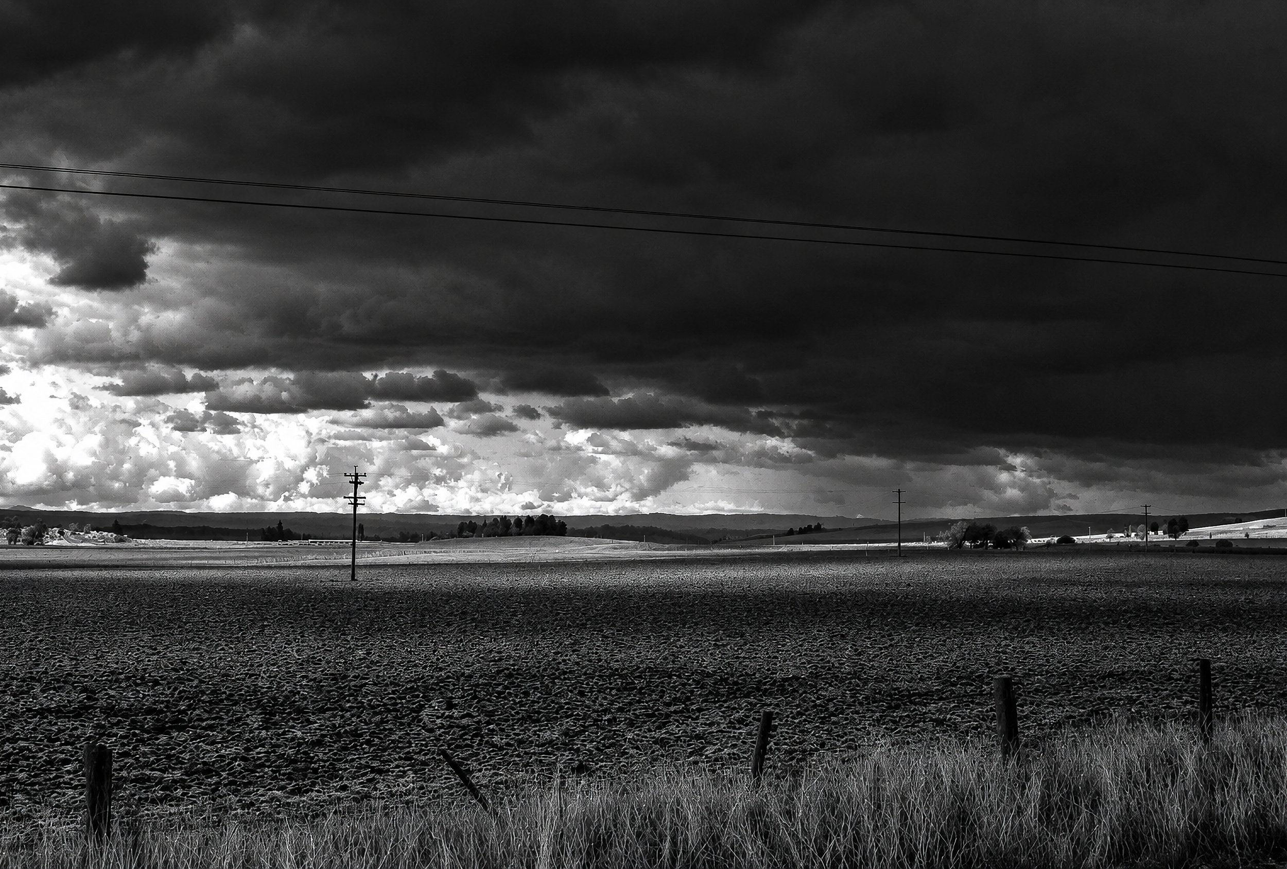 Before the Storm - The Victorian plains stretch towards a distant horizon beneath a vast bank of dark cloud. Power poles and fence lines provide a quiet human scale against the enormous sky, while the fields disappear towards the distant hills.
