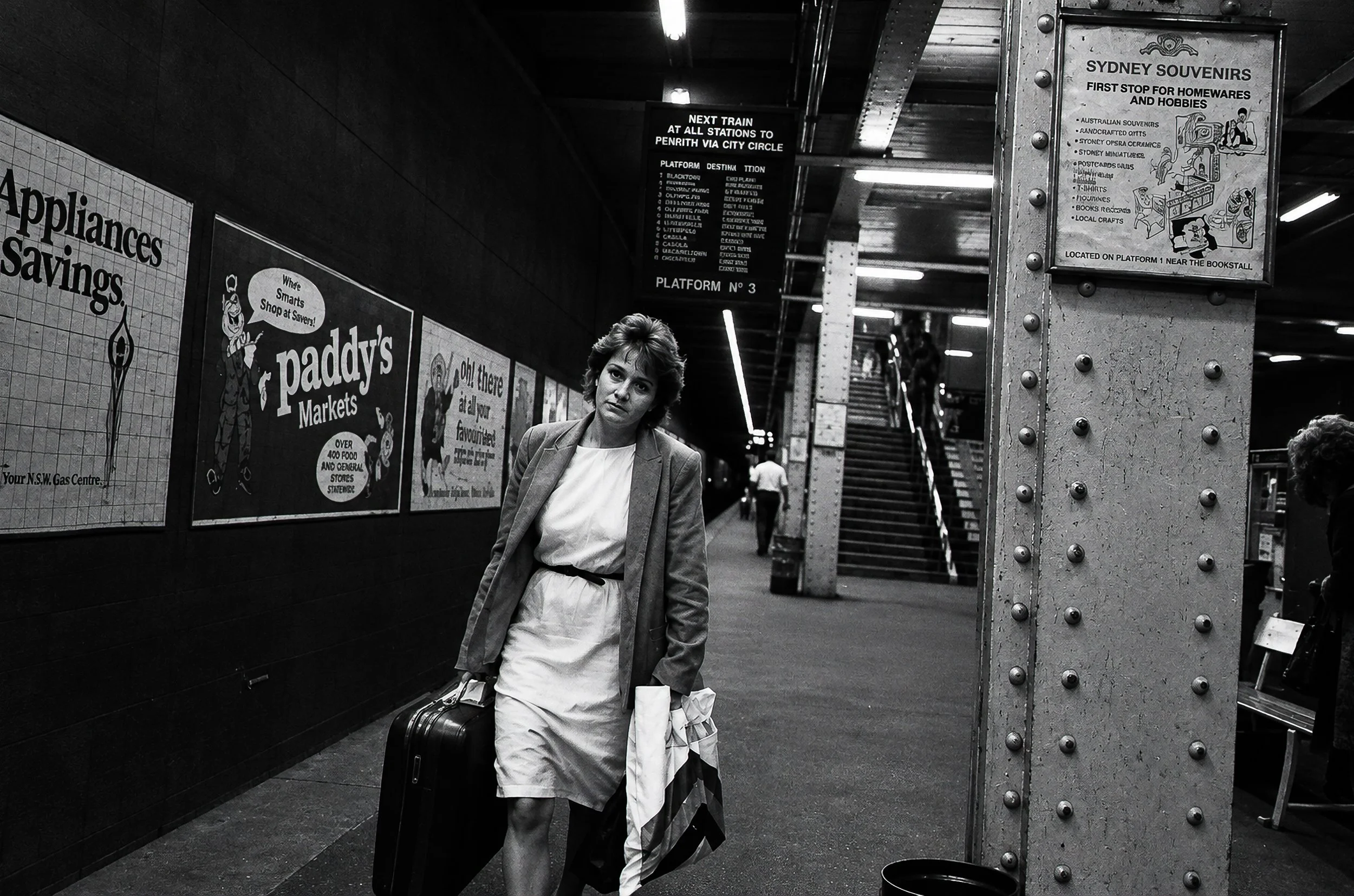Arriving - A traveller carrying bags approaches along the platform, framed by advertisements, station signage and the heavy ironwork of the railway structure. It is an uncomplicated moment of arrival and movement through the city.