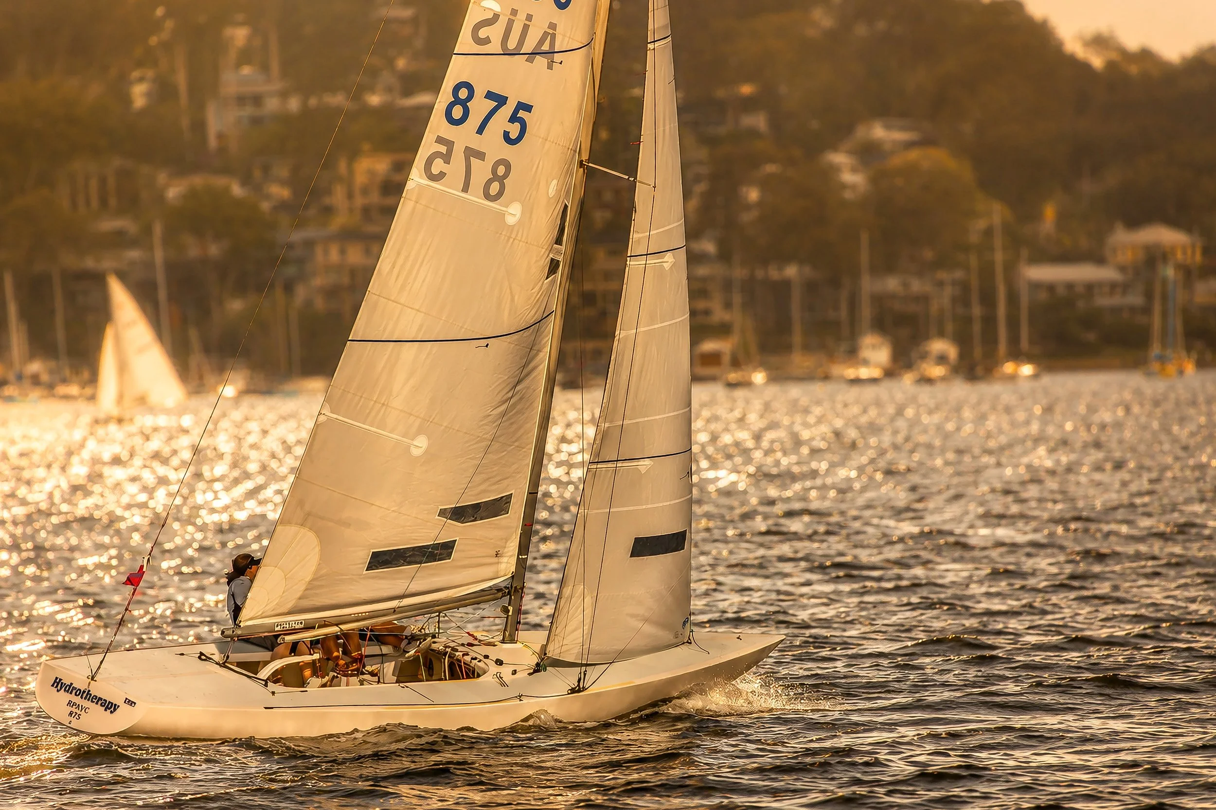 A sailboat racing on a body of water during sunset, with the hull and sails visible and a cityscape with houses and trees in the background.