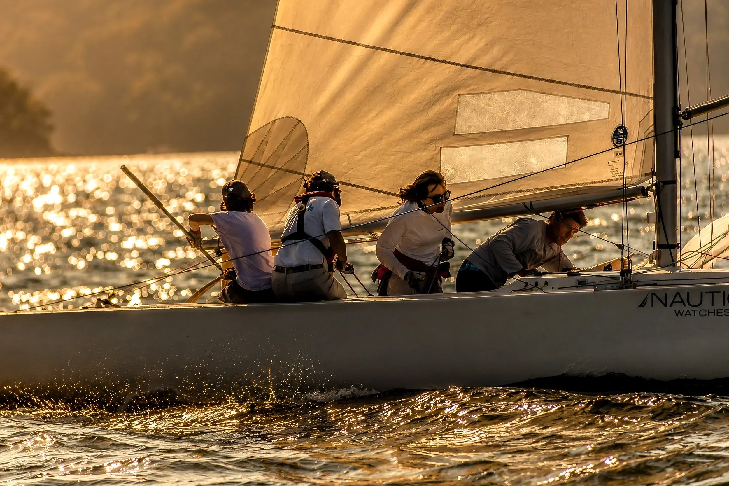 A sailboat with four crew members racing on the water during sunset, with sparkling reflections on the surface.