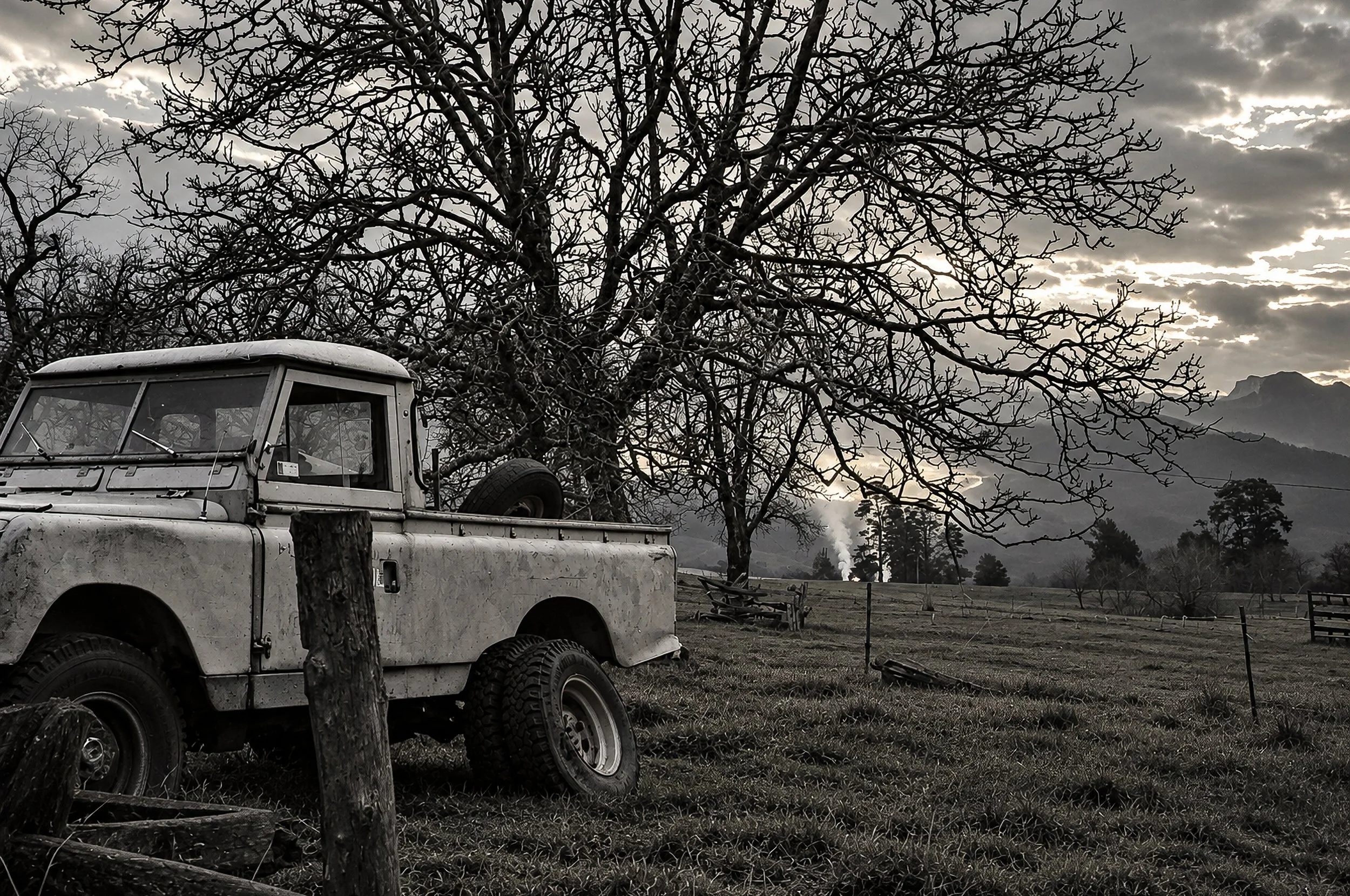 Along the High Country Road - A rugged four-wheel-drive sits beneath a spreading tree beside open farmland, with mountains rising in the distance. The combination of vehicle, country landscape and dramatic sky evokes the isolation and changing charac