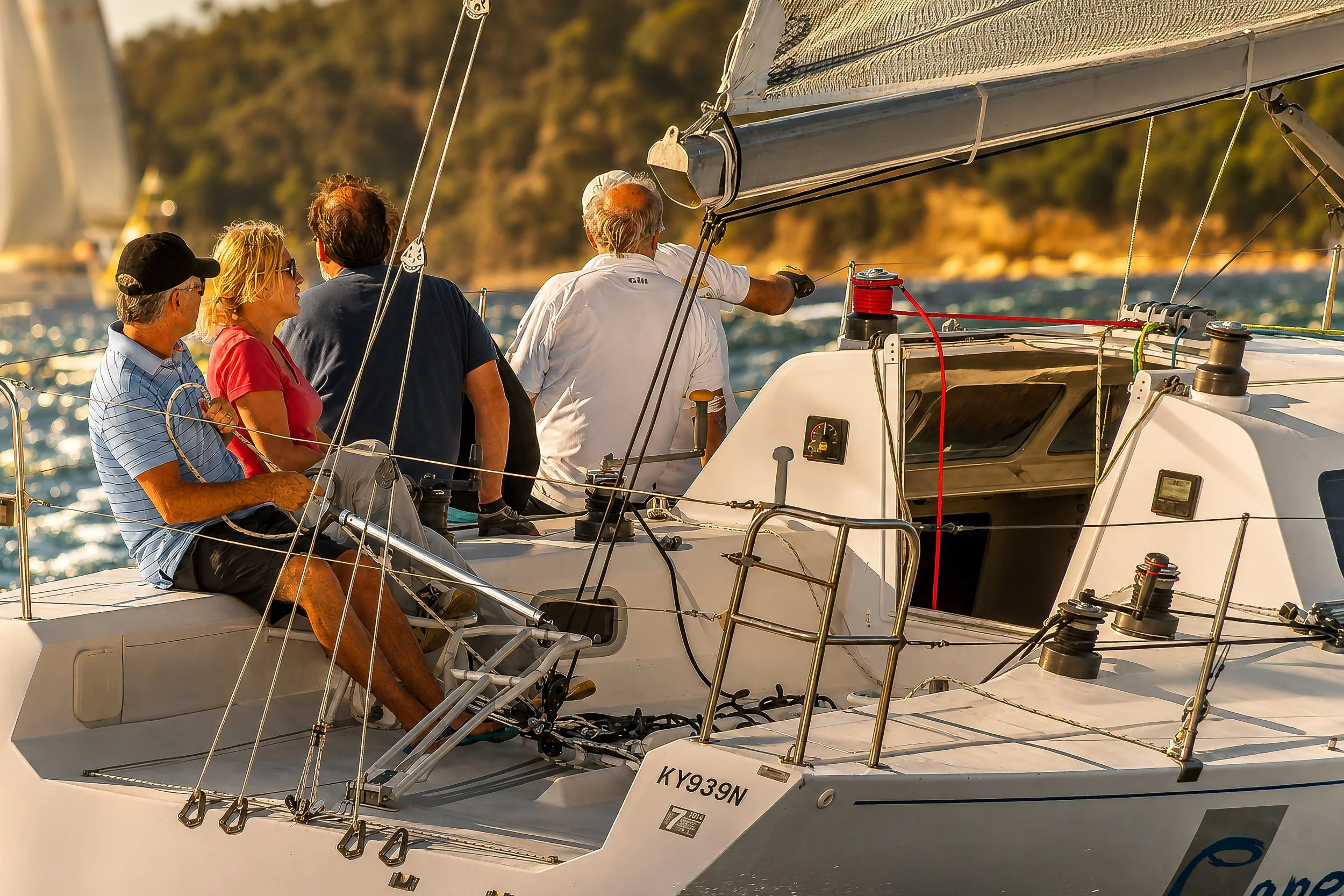 Four people on a sailboat, with a man steering and others seated, sailing on the water with a hilly shoreline in the background during sunset.