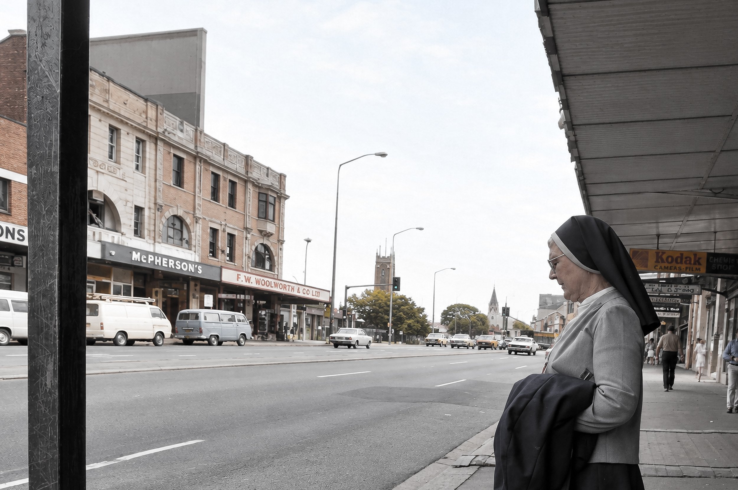 Waiting Beneath the Awning - A nun stands beneath a city awning with the busy Sydney street stretching behind her. The strong architectural lines and contrasting light create a quiet, observational moment within the urban environment.