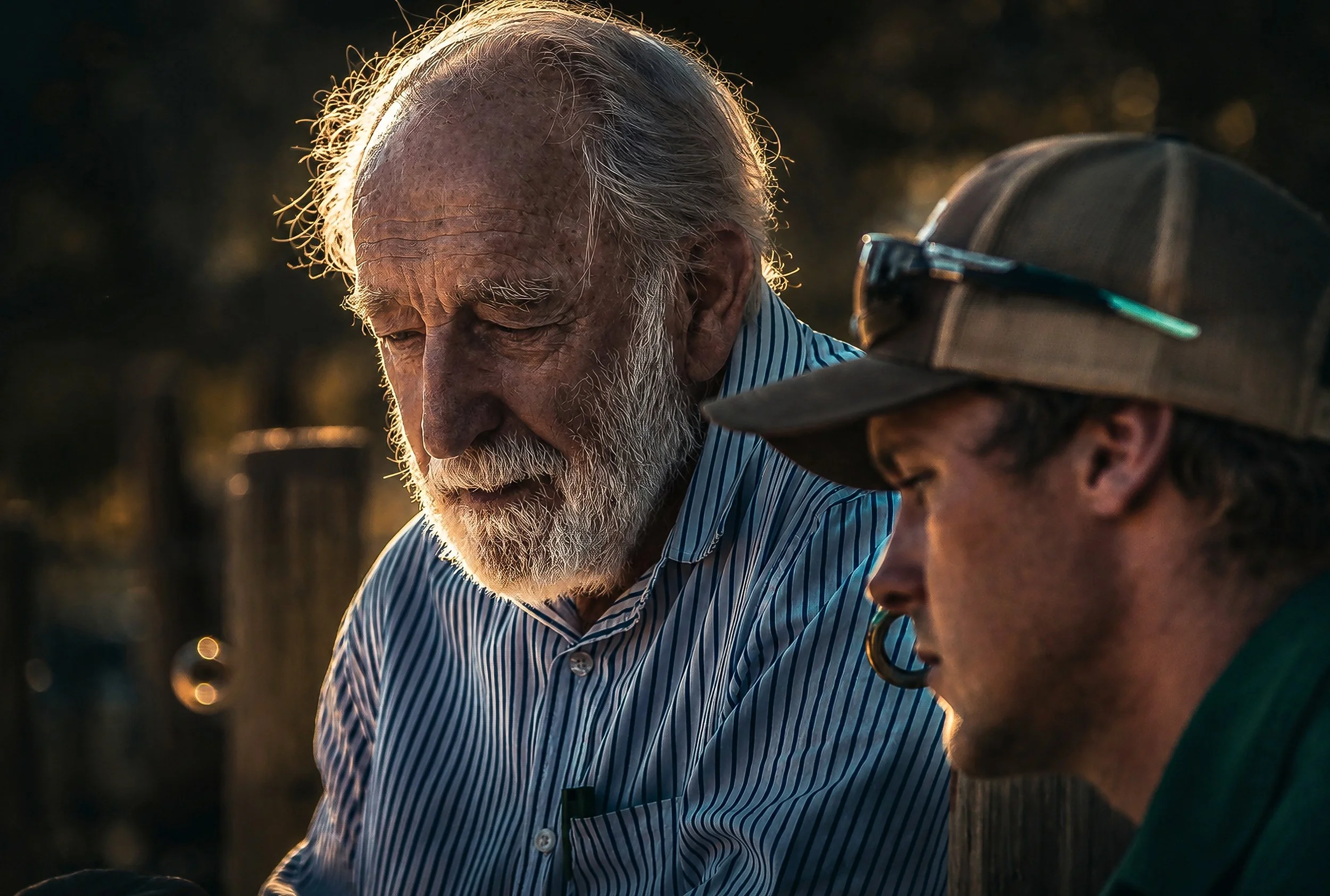 Close-up of two elderly men engaged in conversation outdoors during sunset, with a dark background and warm lighting highlighting their faces.